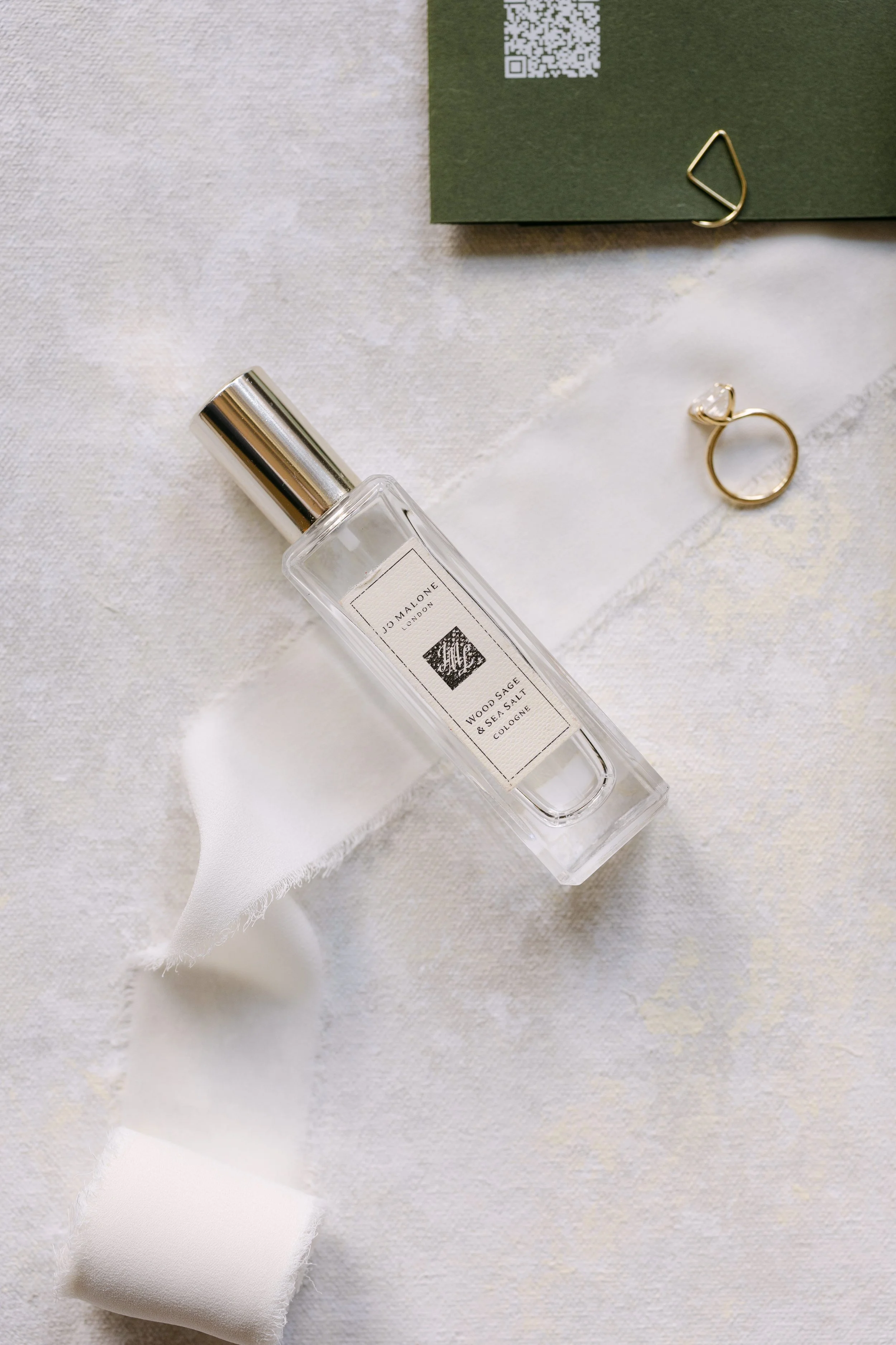 A bottle of Jo Malone Wood Sage & Sea Salt Cologne on a white textured surface, along with a gold ring, a gold paperclip, a green piece of paper with a QR code, and a white cloth ribbon.