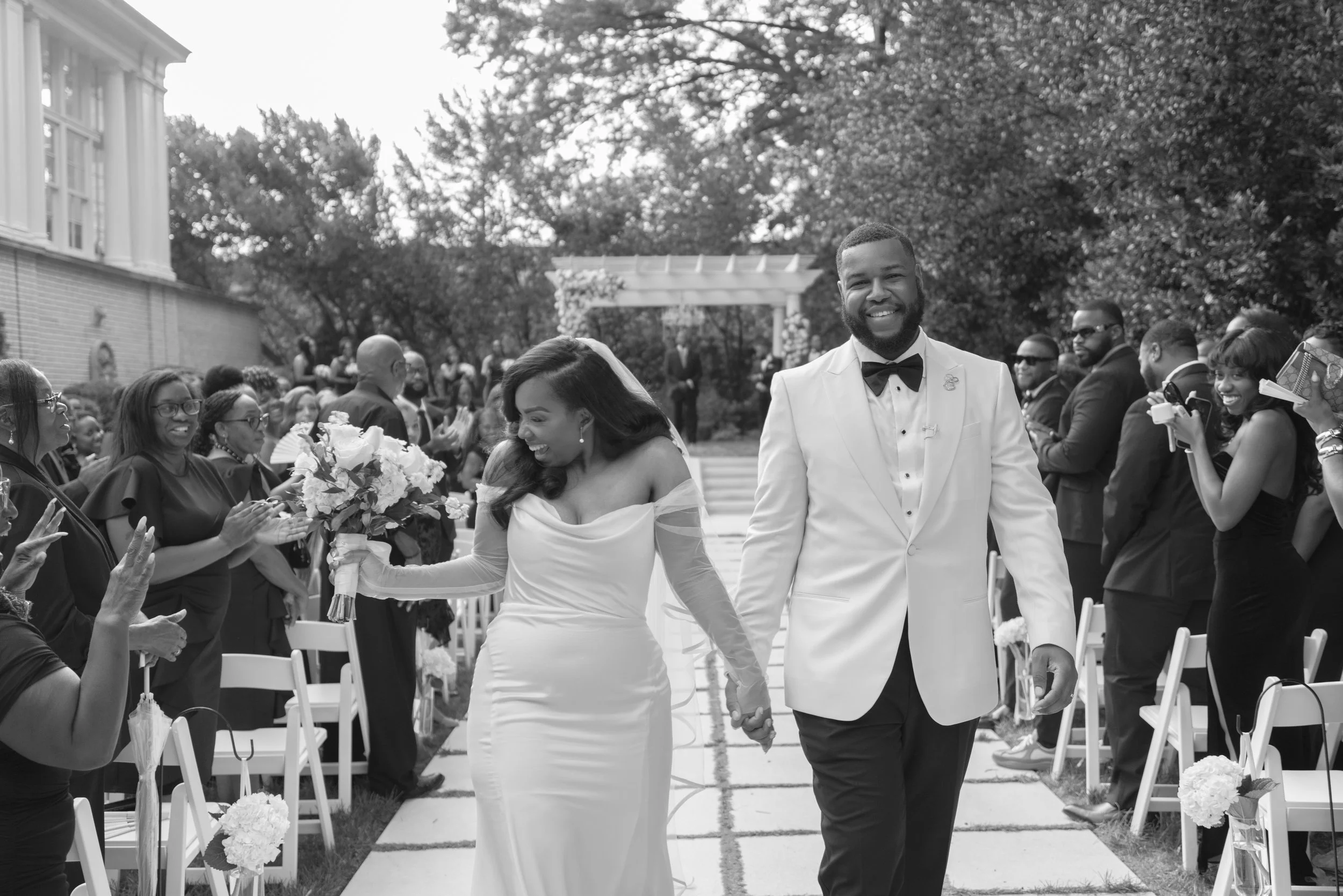 A black-and-white photograph of a wedding ceremony outdoors, with the bride and groom holding hands and smiling as they walk down the aisle. The bride is wearing a white wedding dress and holding a bouquet, while the groom is dressed in a white tuxed