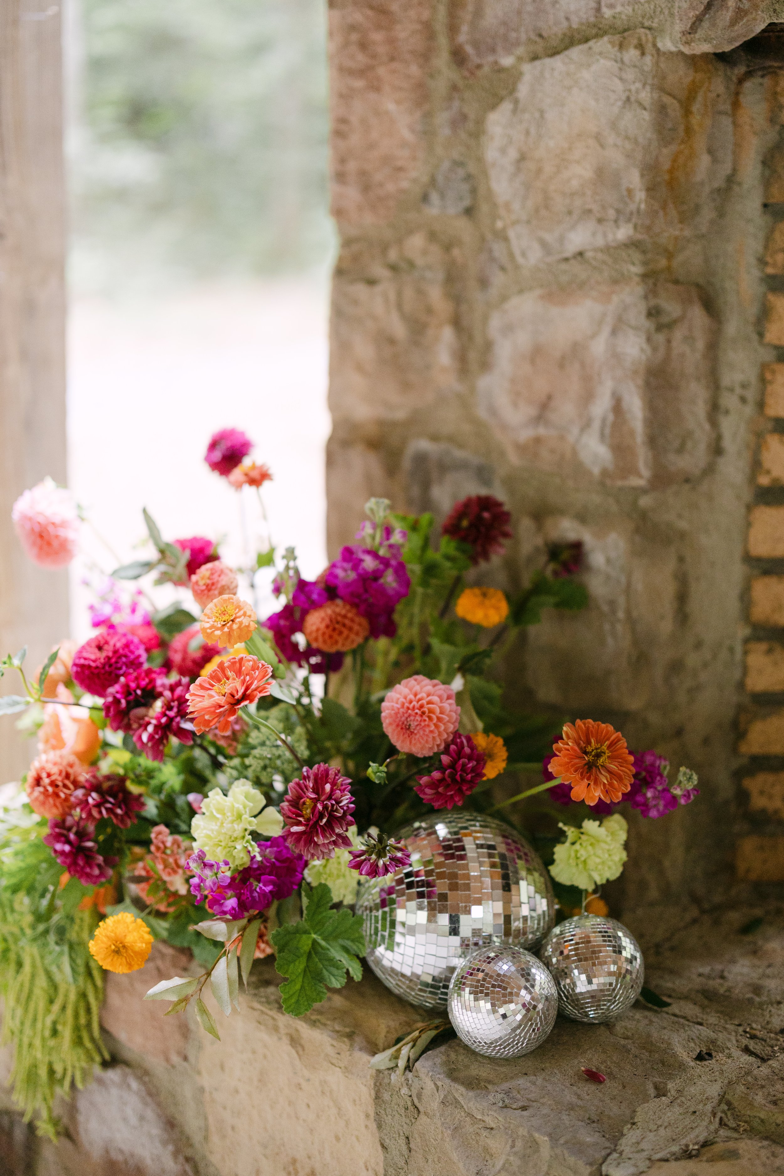 A colorful bouquet of flowers in a silver disco ball vase, placed on a windowsill next to a brick and stone wall.
