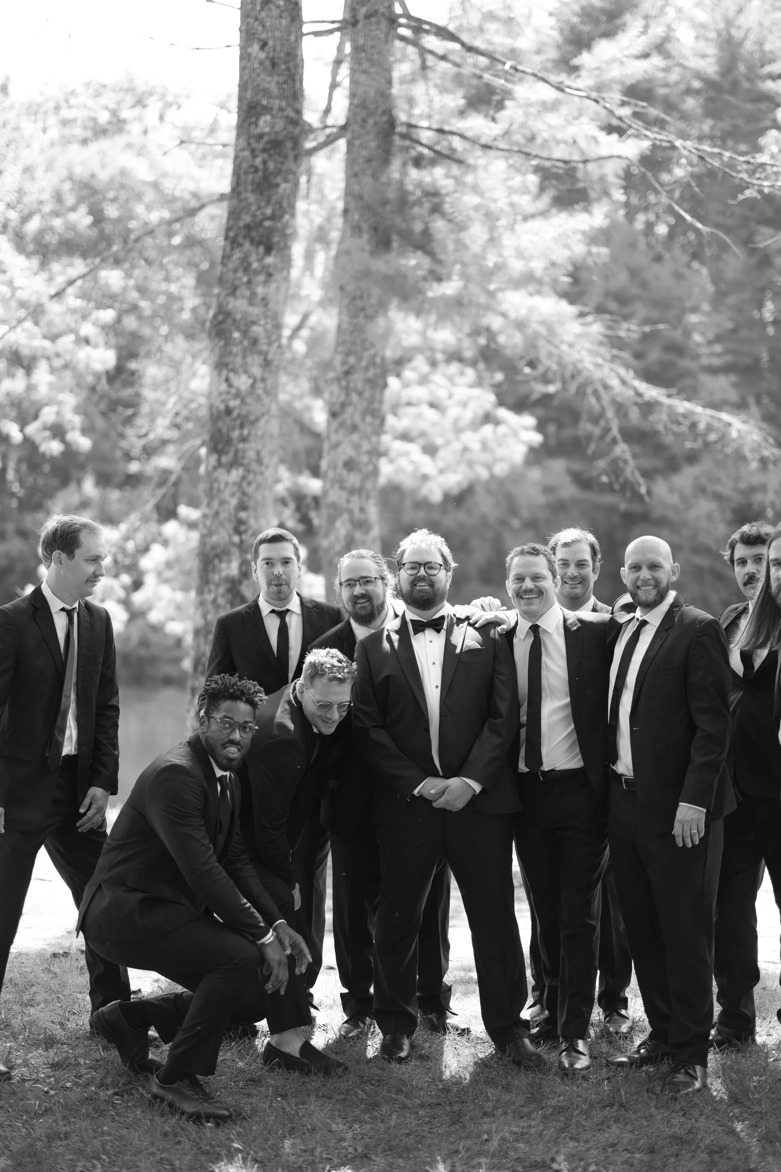 A group of men dressed in suits, posing outdoors in a forested area, smiling and standing close together.