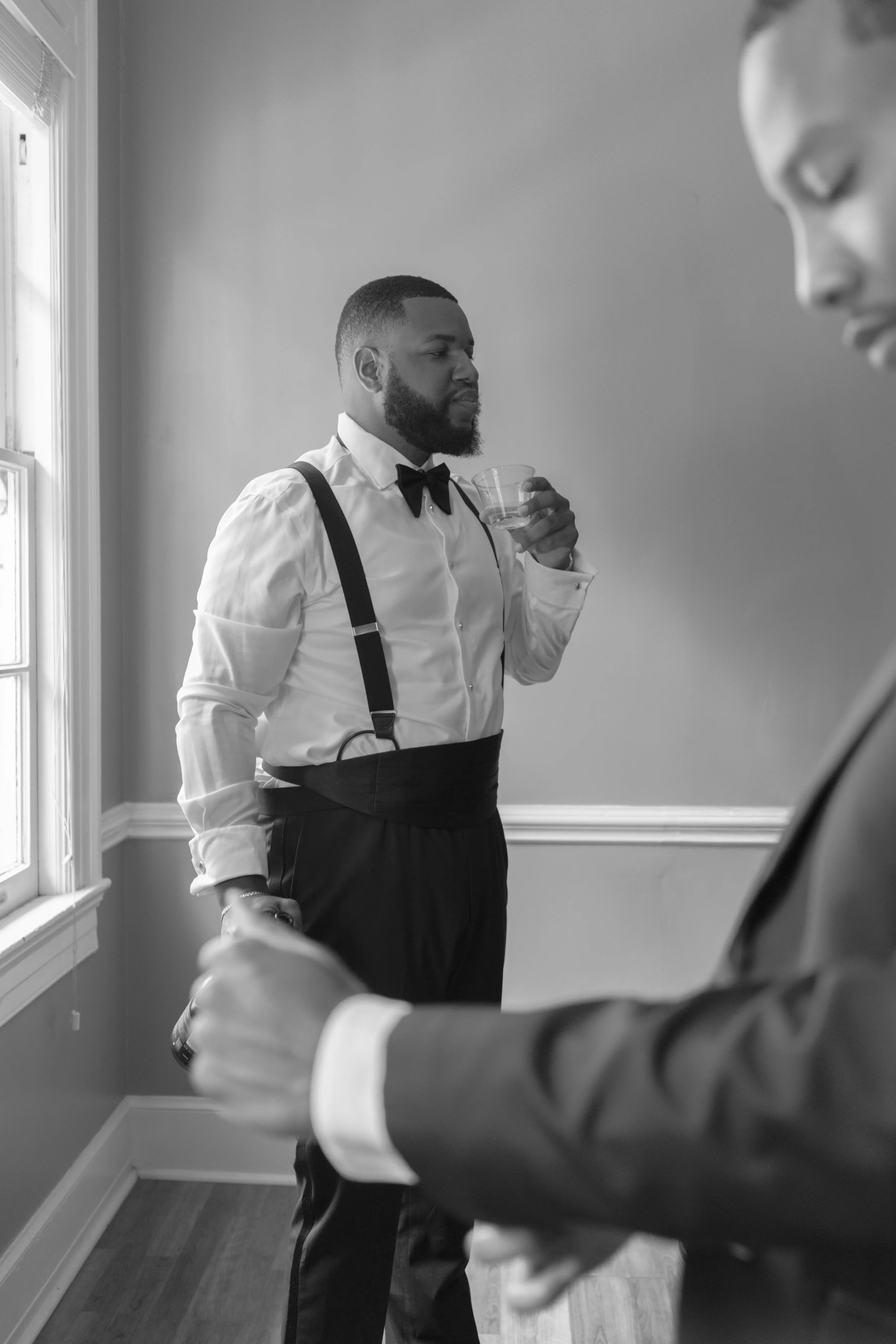 A man with a beard dressed in formal attire including a white shirt, black bow tie, suspenders, and black pants, standing indoors near a window, holding a glass of water, and looking at another person whose shoulder and side of the face are visible i