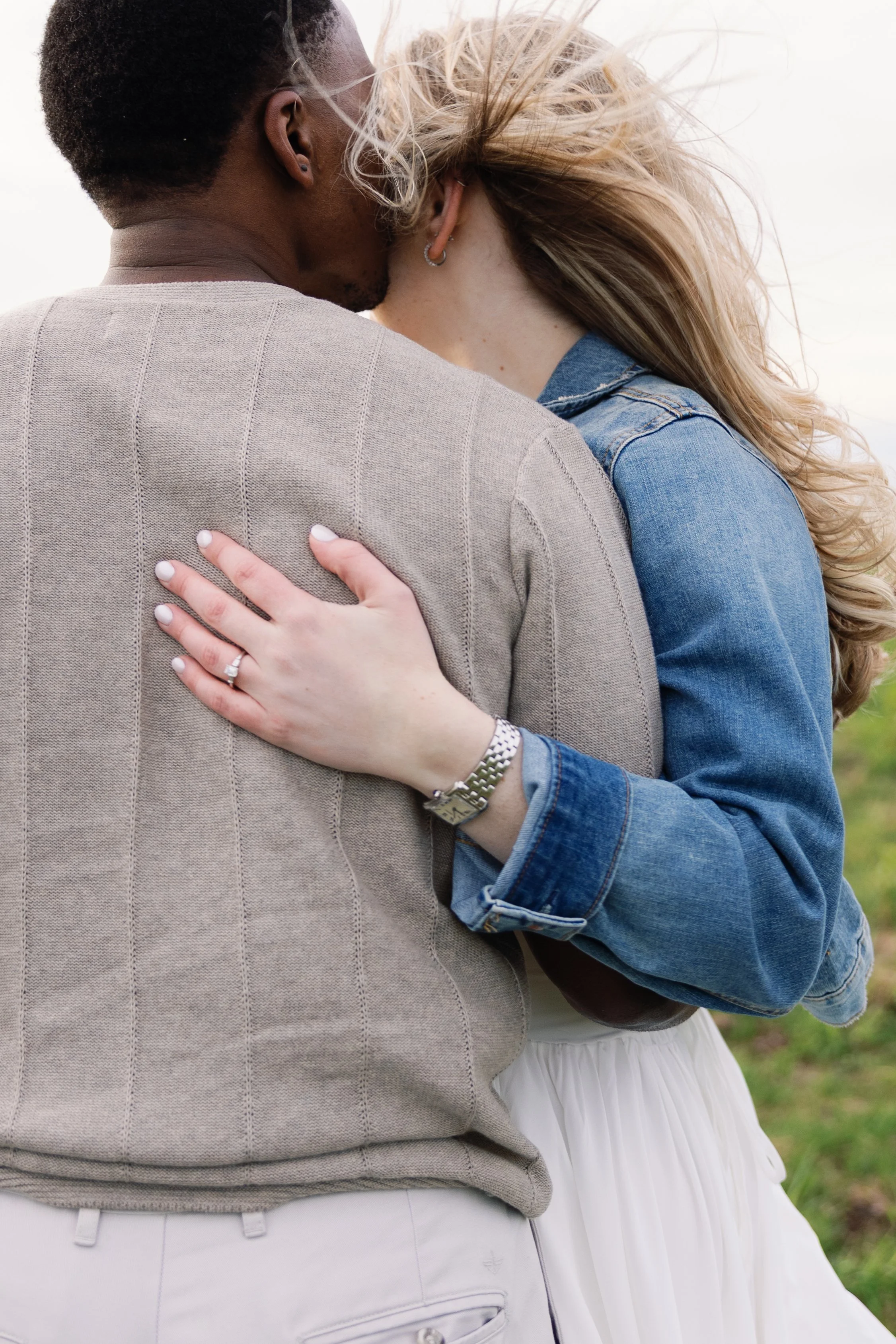 A couple hugging outdoors, focused on their torsos and arms, with a blurred grassy background.