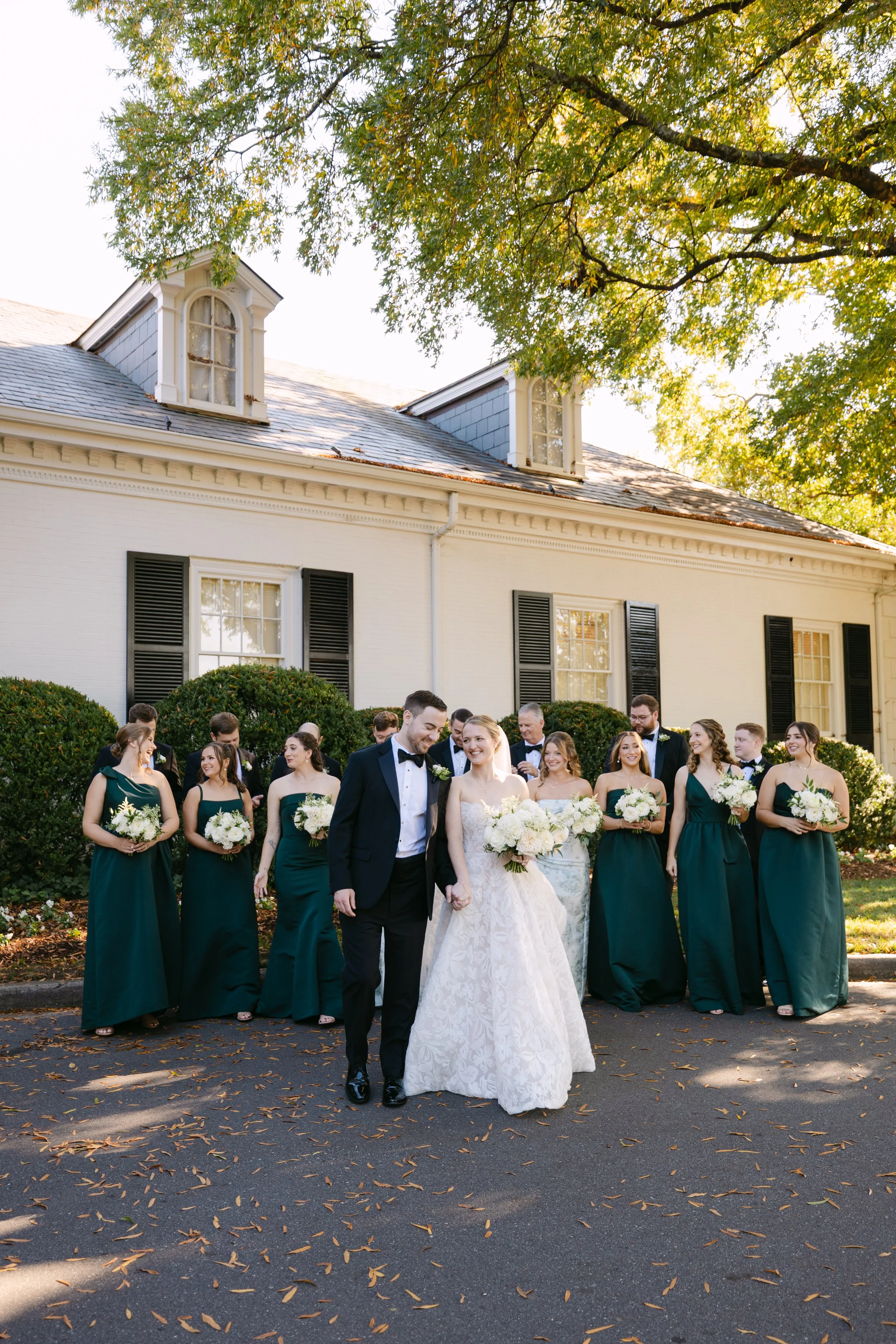 Bride and groom walking together in front of wedding party, outdoors, in front of white house with black shutters, surrounded by greenery, on a sunny day.