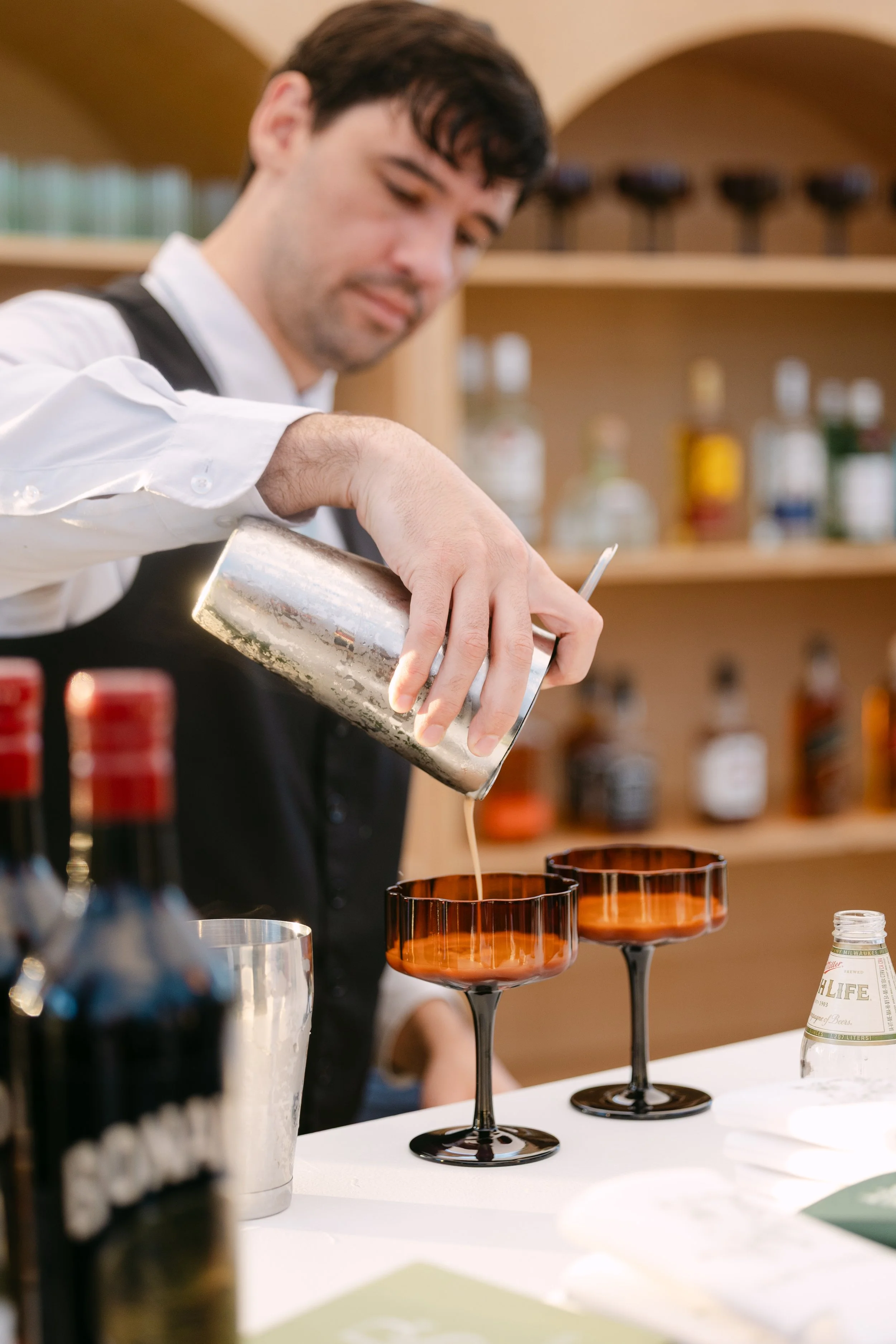 A bartender pouring a drink into glasses at a bar with shelves of liquor bottles in the background.