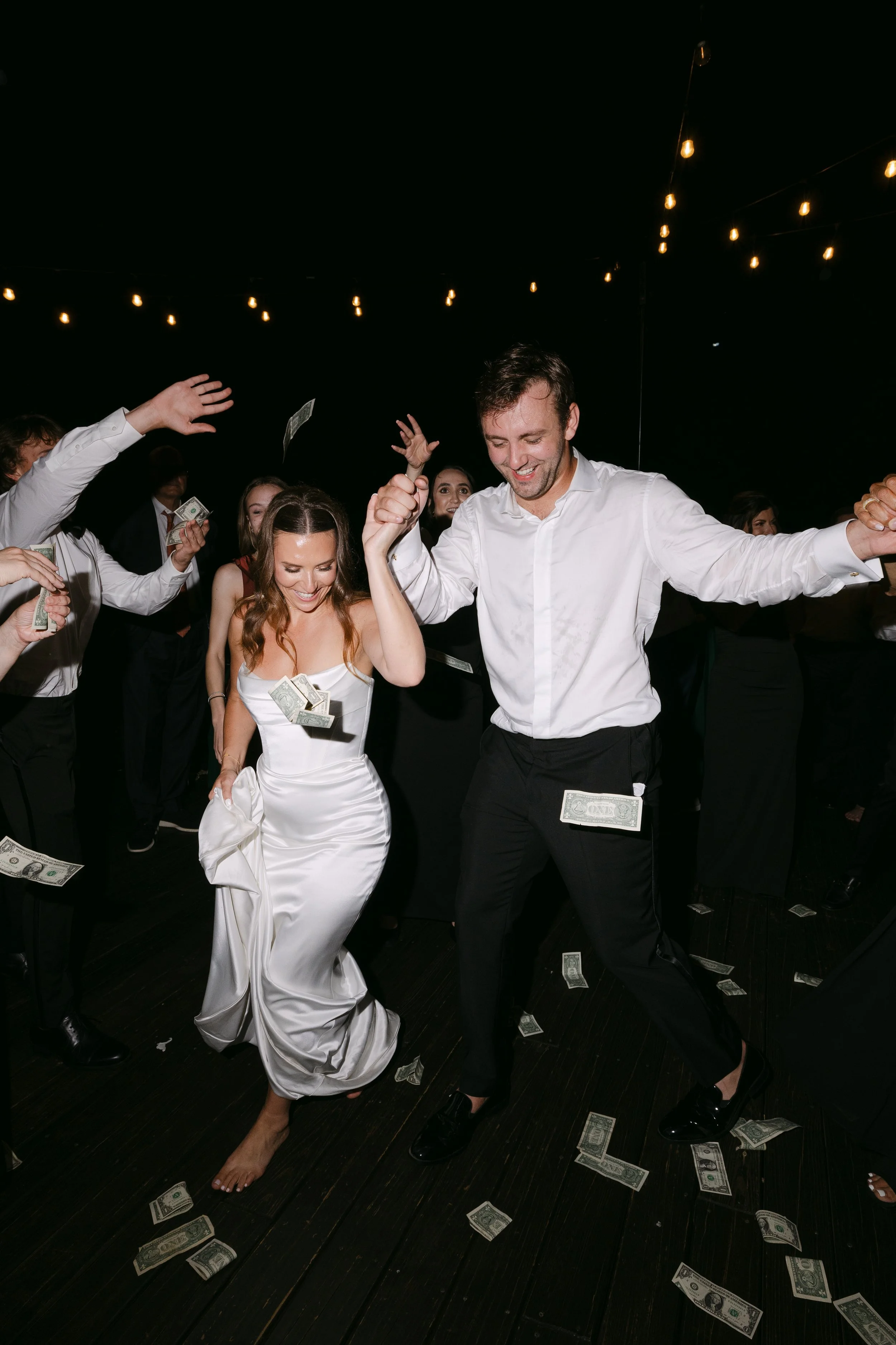 Couple dancing at a celebration, surrounded by guests throwing dollar bills, with string lights overhead.