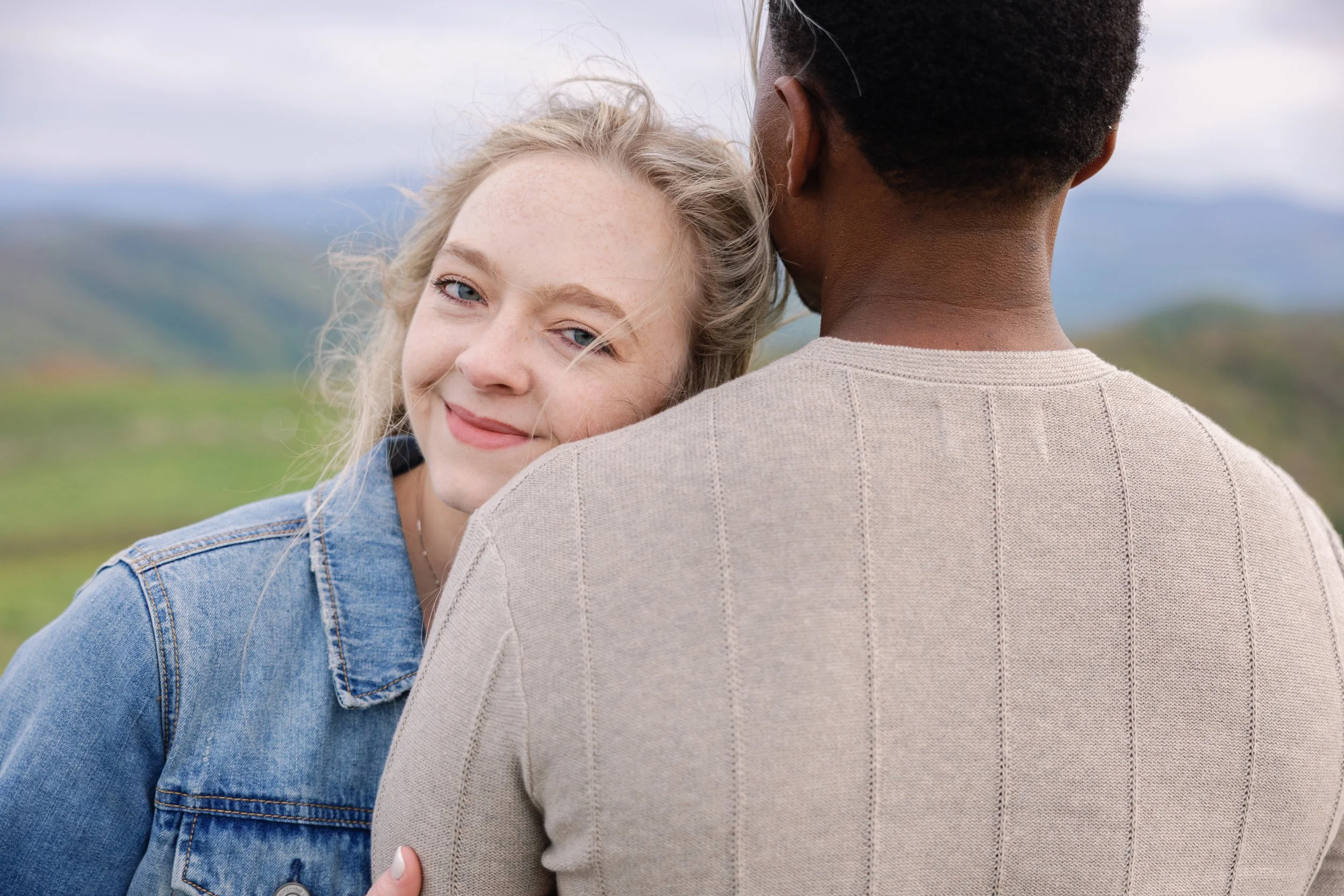 A young woman with blonde hair and blue eyes hugging a man with dark skin and short curly hair outdoors with a cloudy sky in the background.