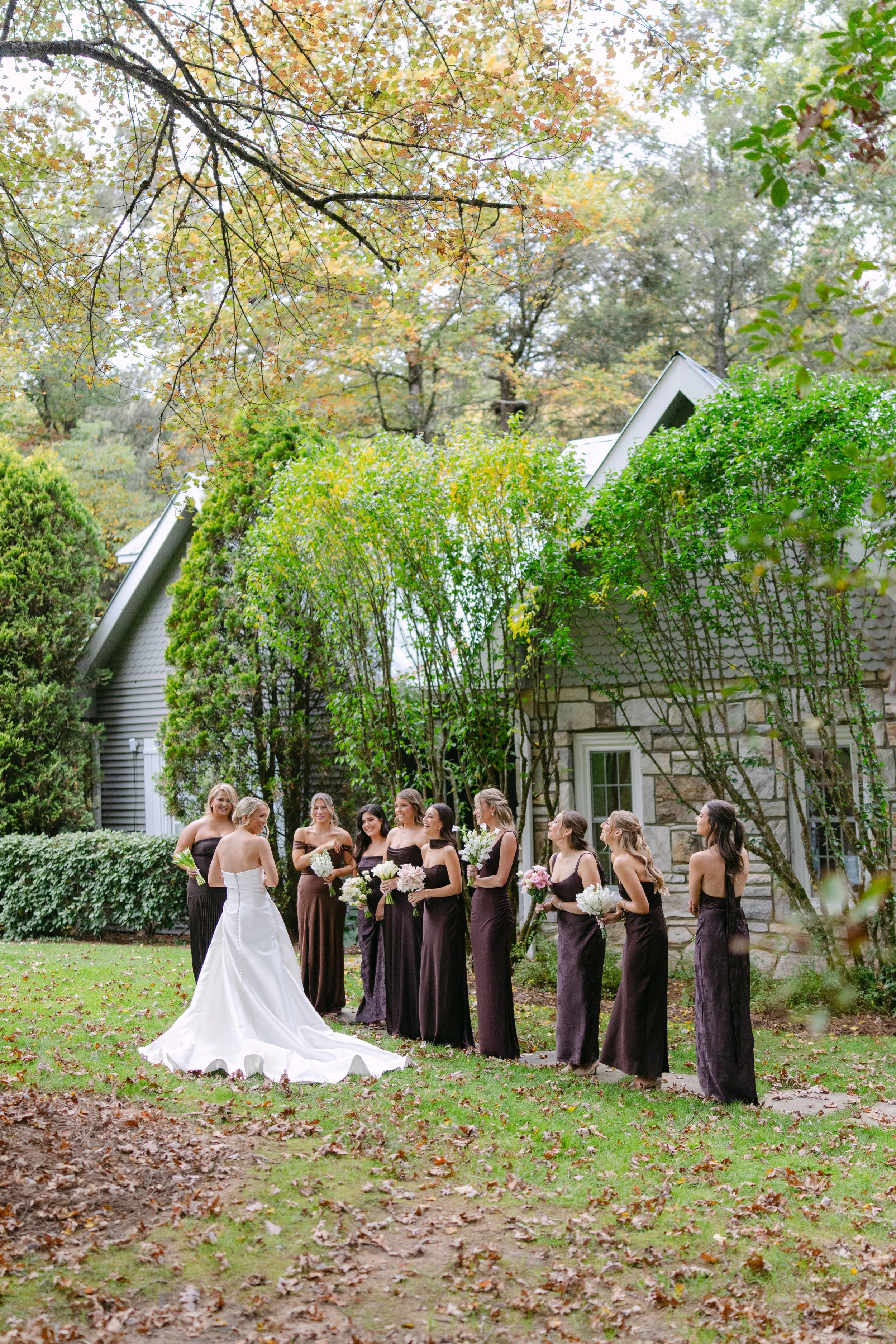 Bride in a white wedding gown standing with bridesmaids in dark dresses and bouquets outside a stone house in an autumn garden