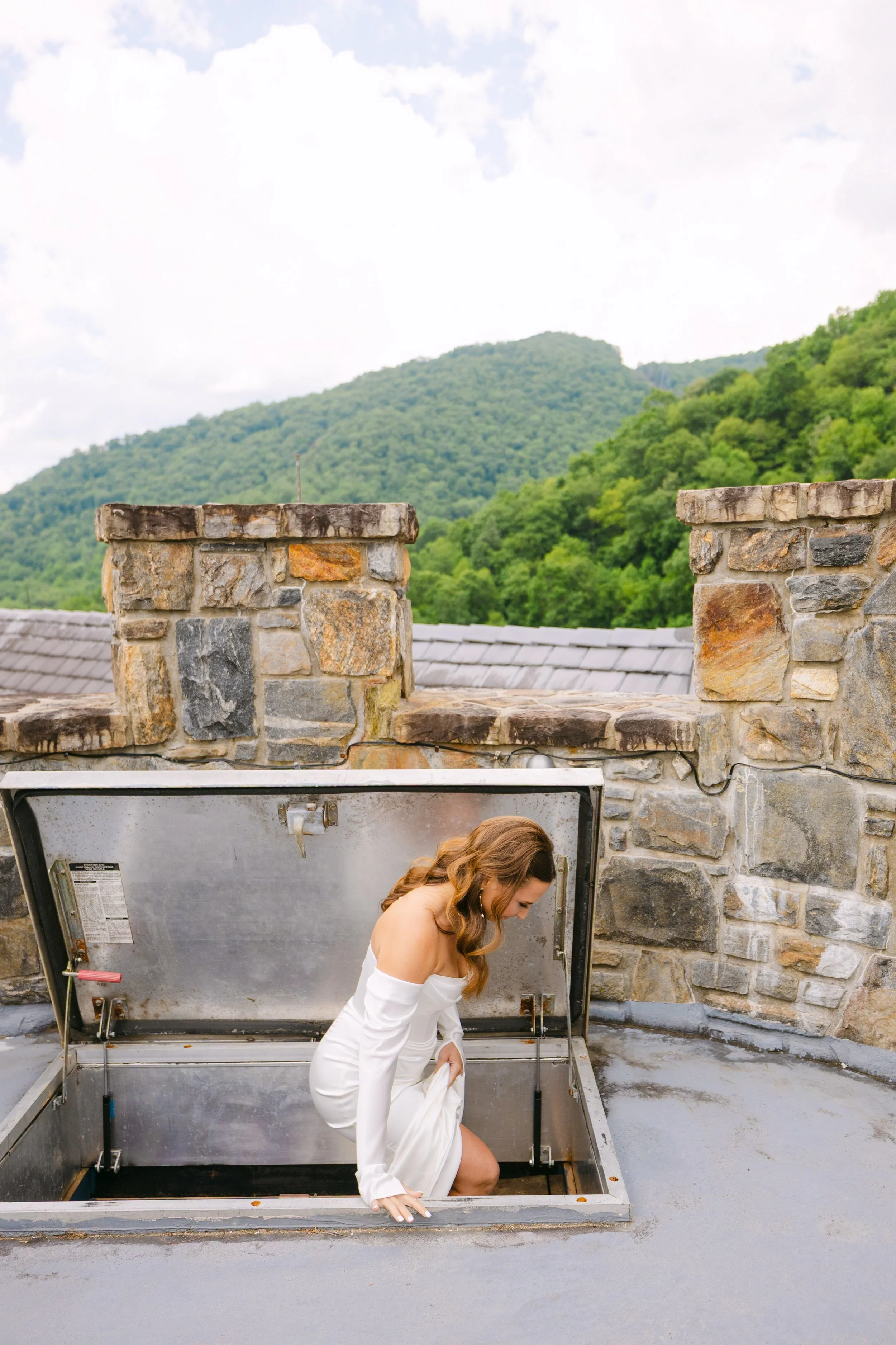 Woman in white dress climbing out of a rooftop attic vent in front of green hills and cloudy sky
