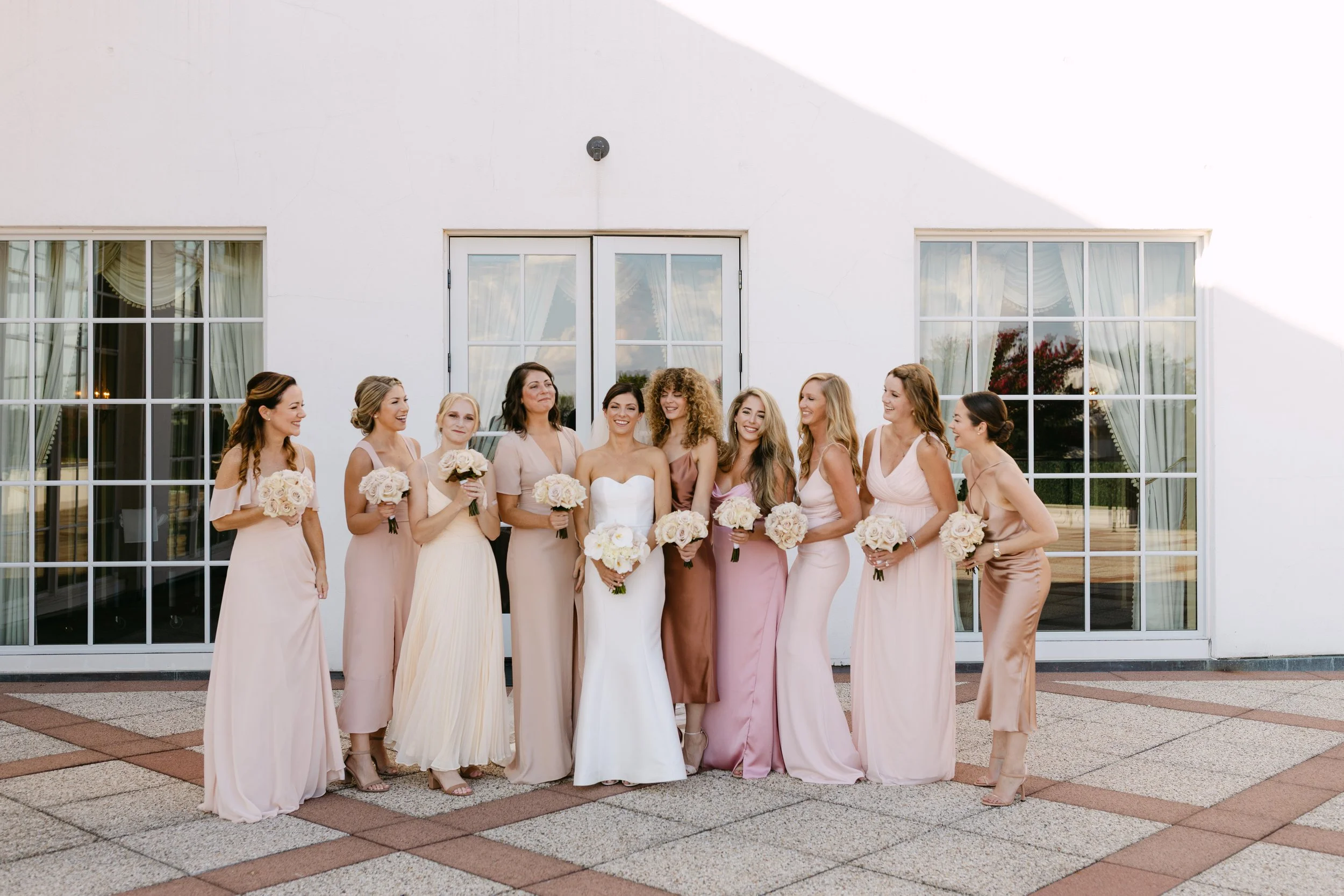 A group of eleven women, including a bride in a white wedding dress, standing outdoors in front of a large white building with glass-paneled doors and windows, all holding bouquets of white roses.