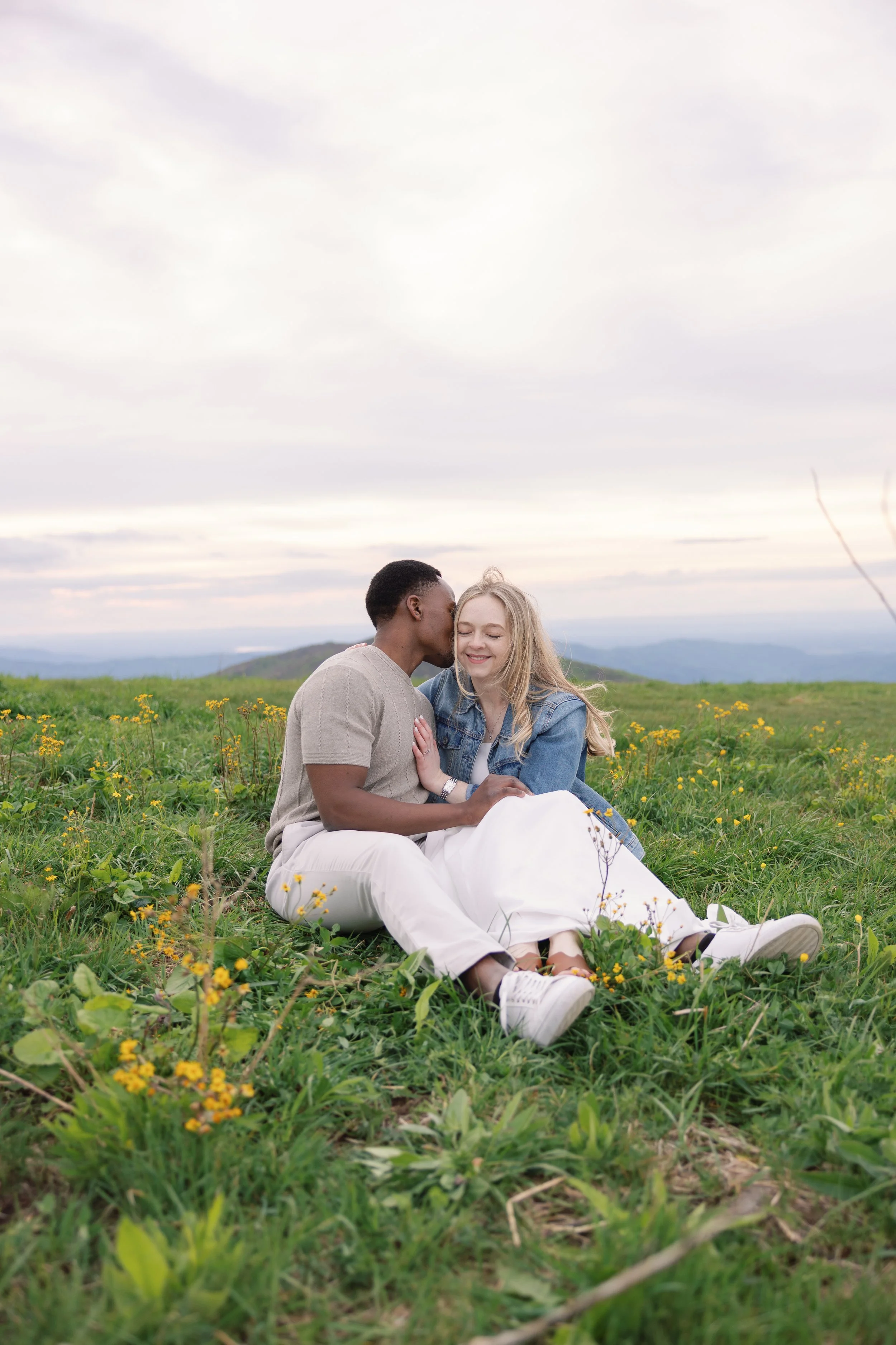 A couple sitting happily in a field of green grass and yellow flowers with a scenic mountain and cloudy sky background.