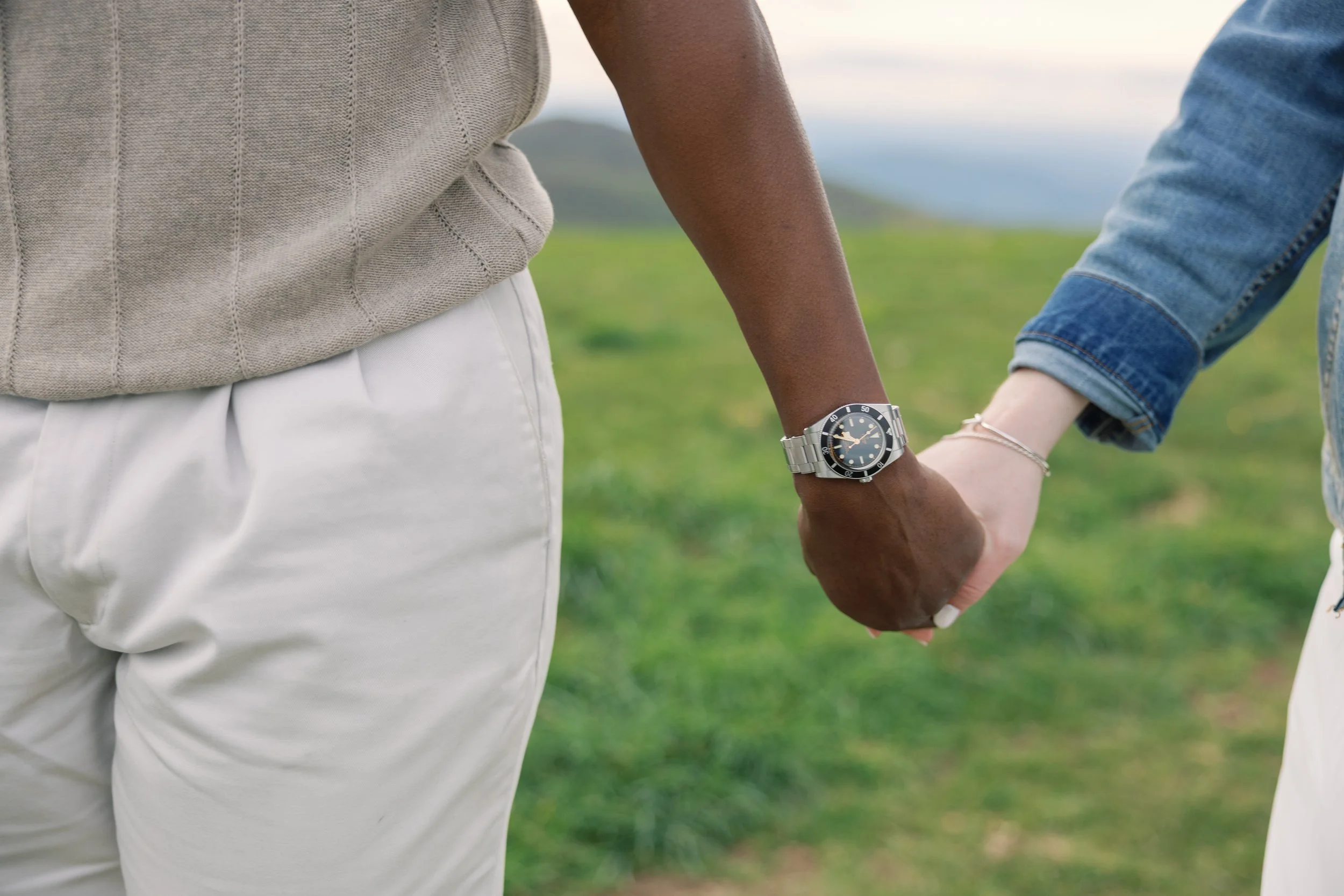 Close-up of two people holding hands outdoors, with one person wearing a silver watch and casual clothing, and the background shows a grassy field.