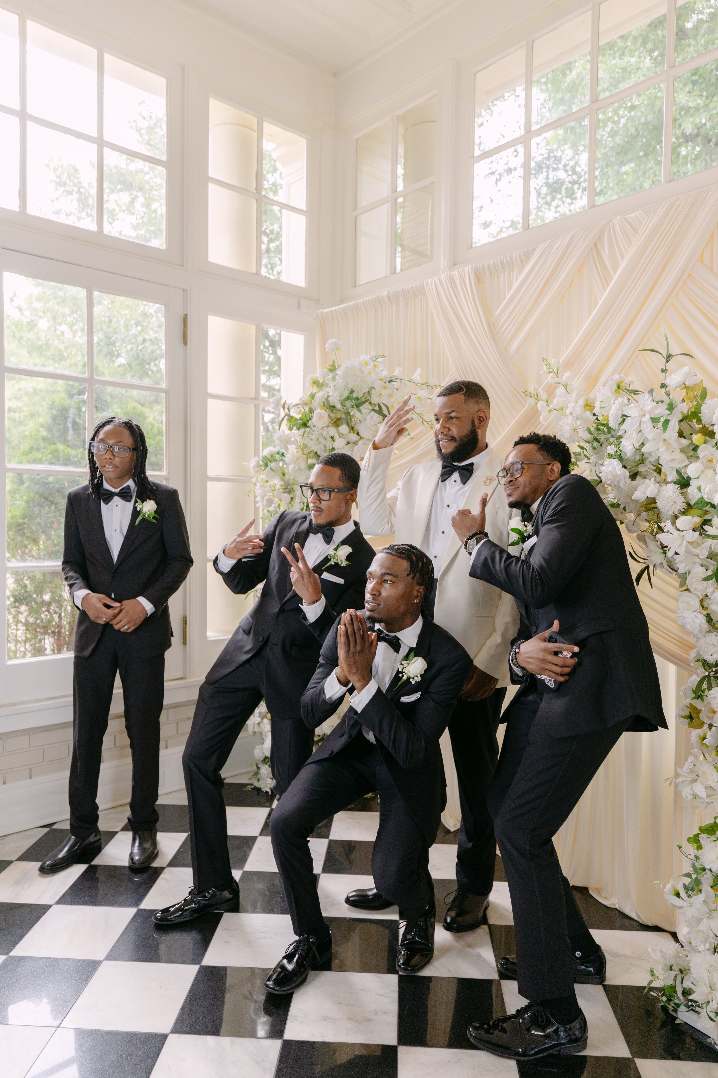 A group of men and a woman dressed in tuxedos and formal wear at a wedding ceremony, standing near a decorated backdrop with white flowers and cream-colored drapery.