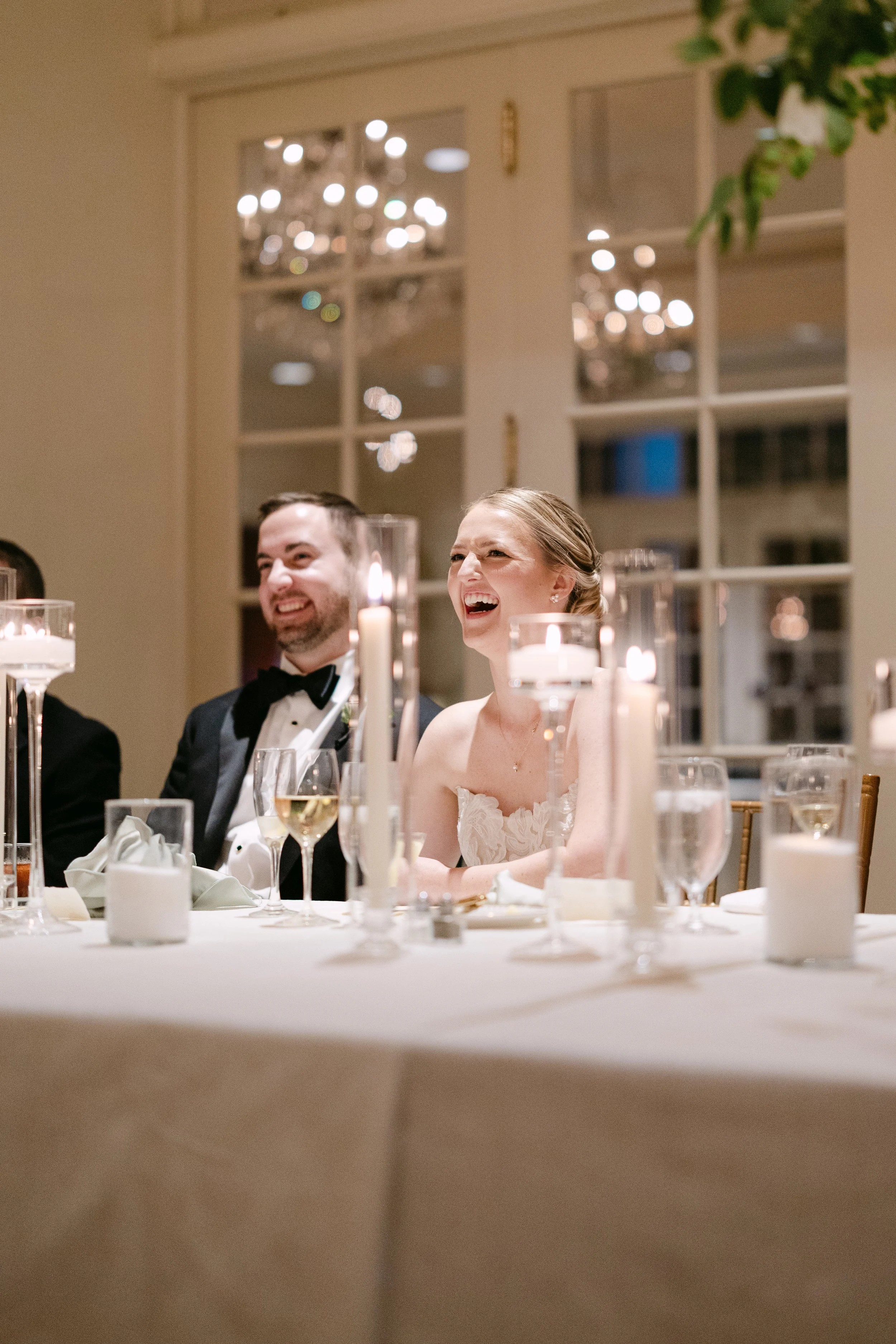 People at a wedding reception sitting at a table with candles and glasses, smiling and laughing.