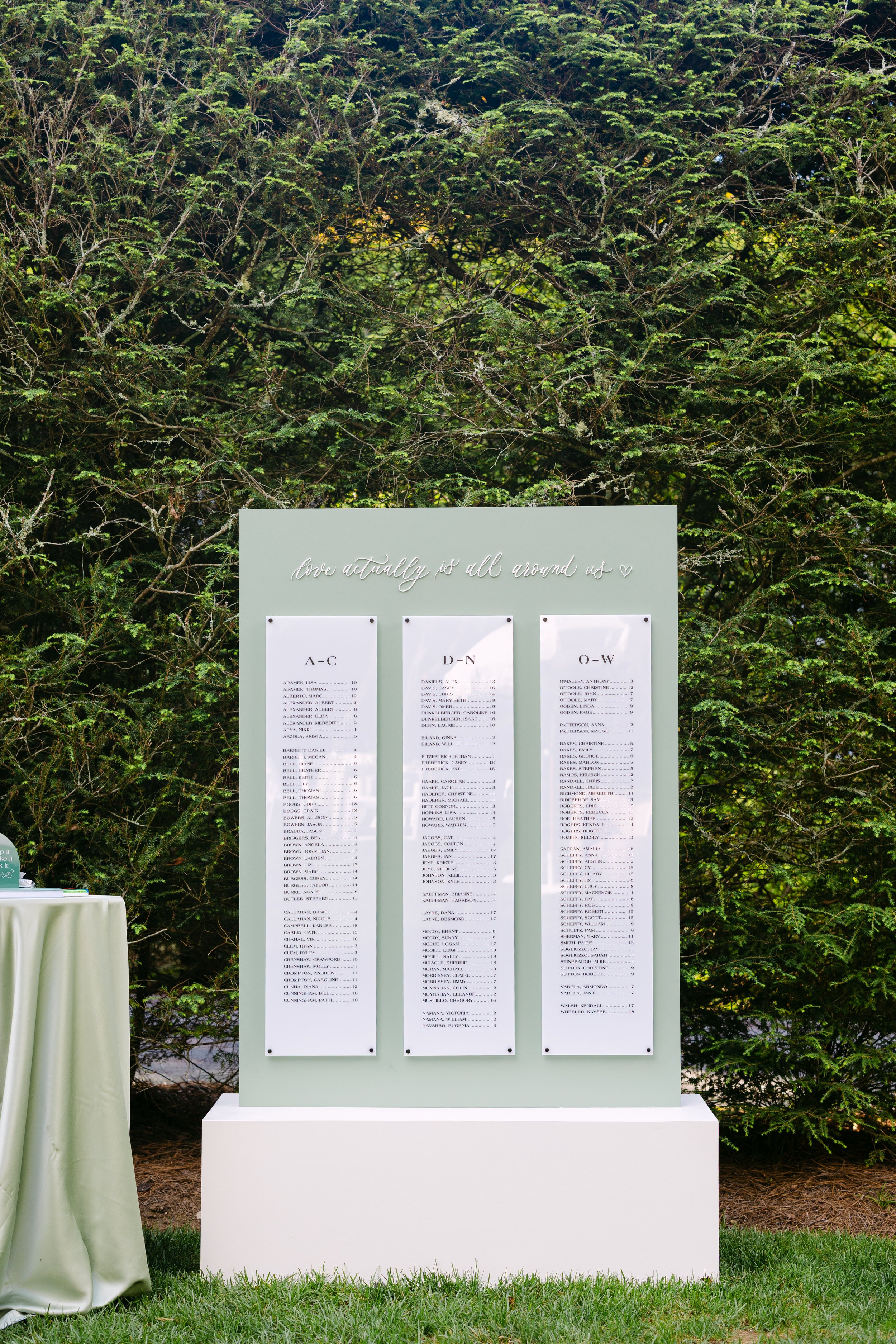 A wedding seating chart displayed on a white board outdoors, with three columns of names organized alphabetically and a decorative sign above that reads "love actually is all around us."