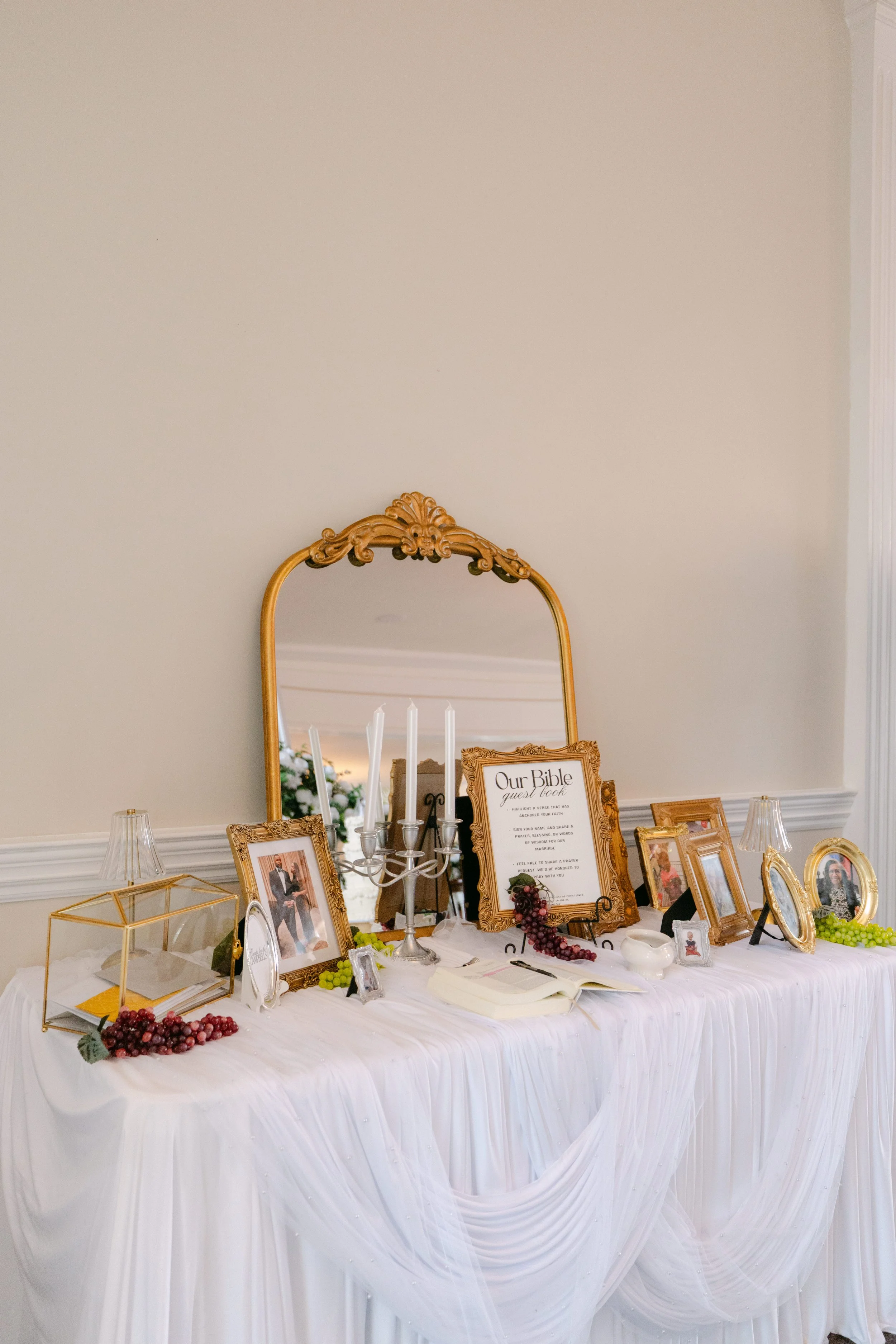 A decorated table with framed photographs, candles, and a sign titled 'Our Bible guest book' at a wedding or celebration event, with a large ornate mirror in the background.