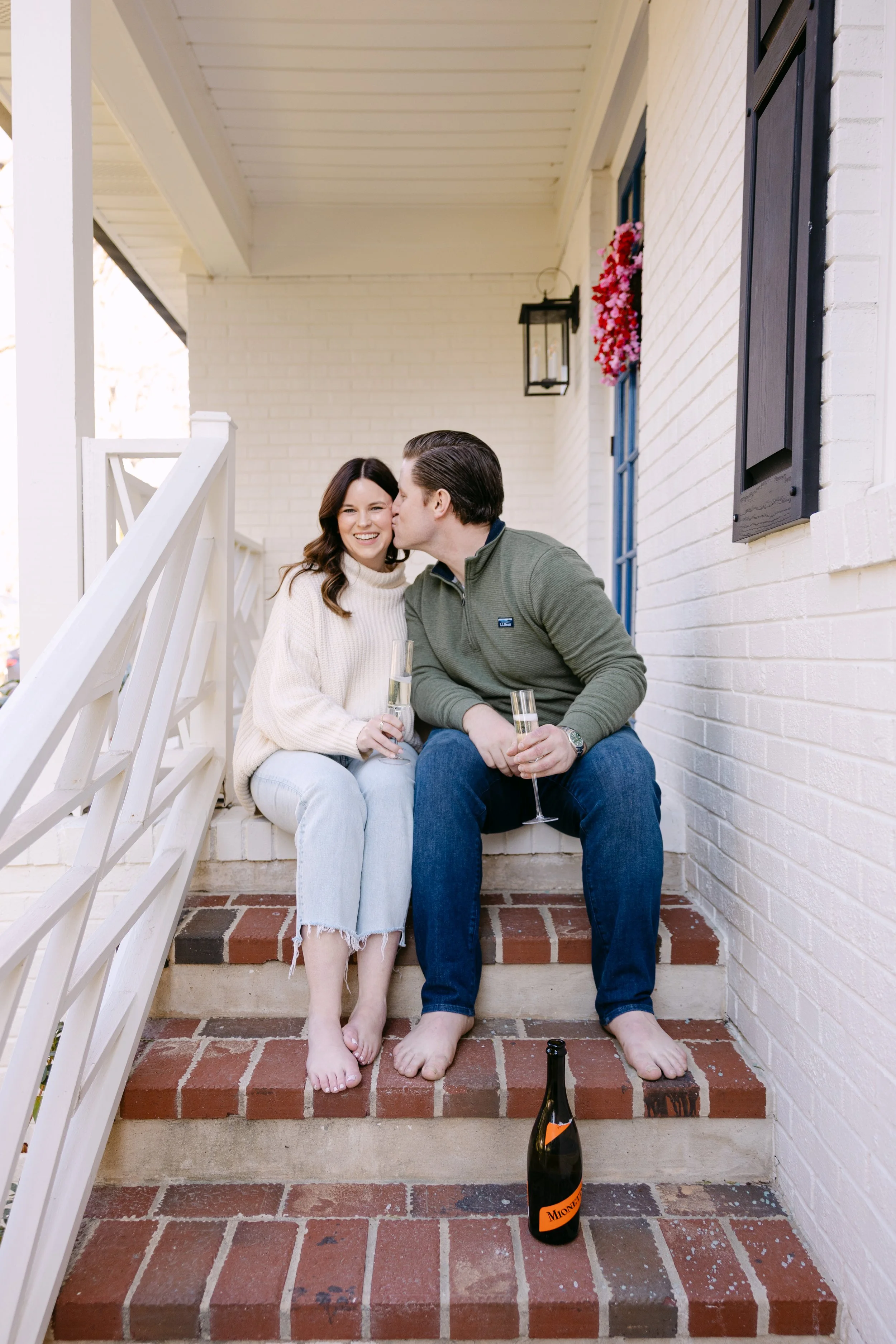 A man and woman sitting on a brick porch steps, sharing a kiss and smiling, holding champagne glasses, with a bottle of champagne on the ground in front of them.