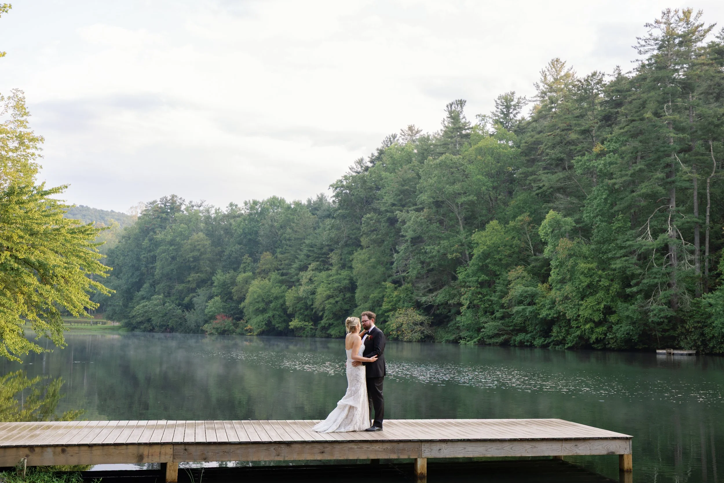 A bride and groom standing on a wooden dock by a lake, surrounded by green trees with a cloudy sky overhead.