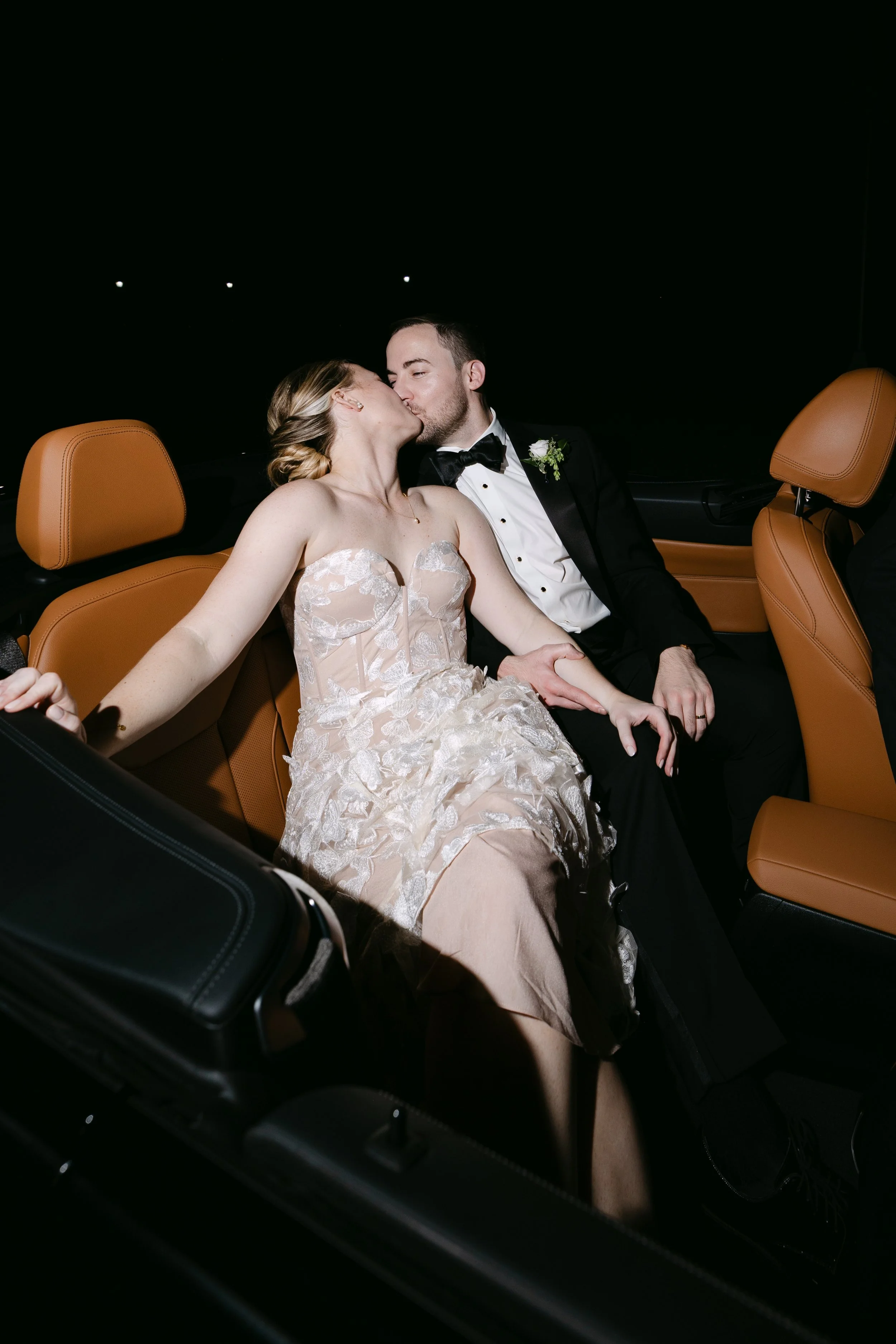 A newlywed couple sharing a kiss in the back seat of a car at night, dressed in wedding attire, with the bride wearing a lace wedding gown and the groom in a tuxedo.
