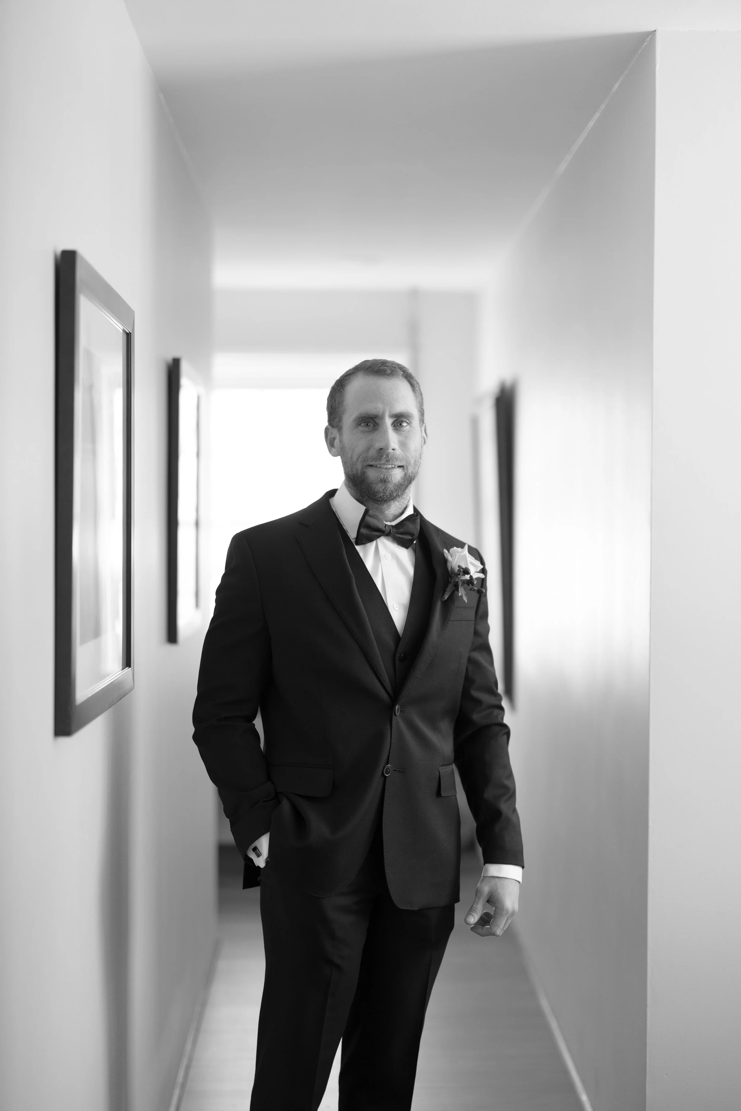 A black-and-white photo of a man dressed in a tuxedo with a bow tie and boutonniere, standing in a hallway with framed pictures on the walls.