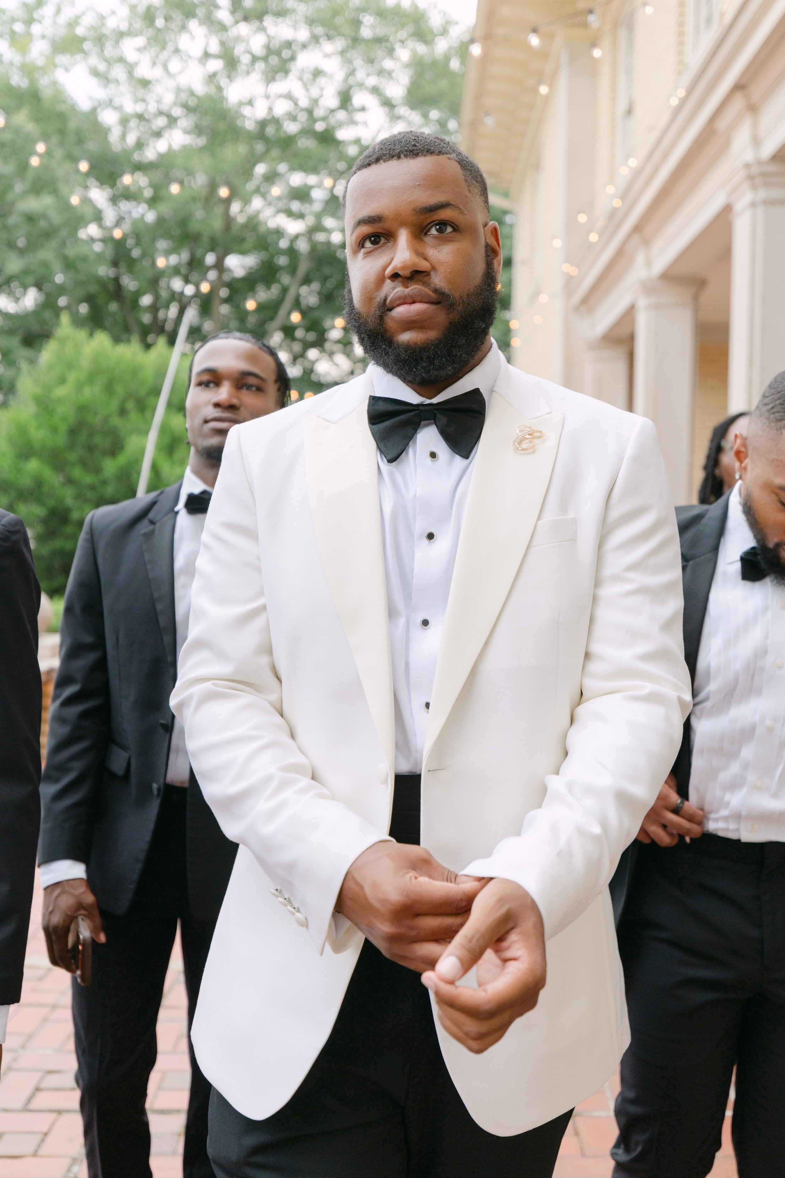 A man wearing a white tuxedo jacket with a black bow tie and a gold brooch, standing outdoors with other men in tuxedos, blurred background of trees and a building with string lights.