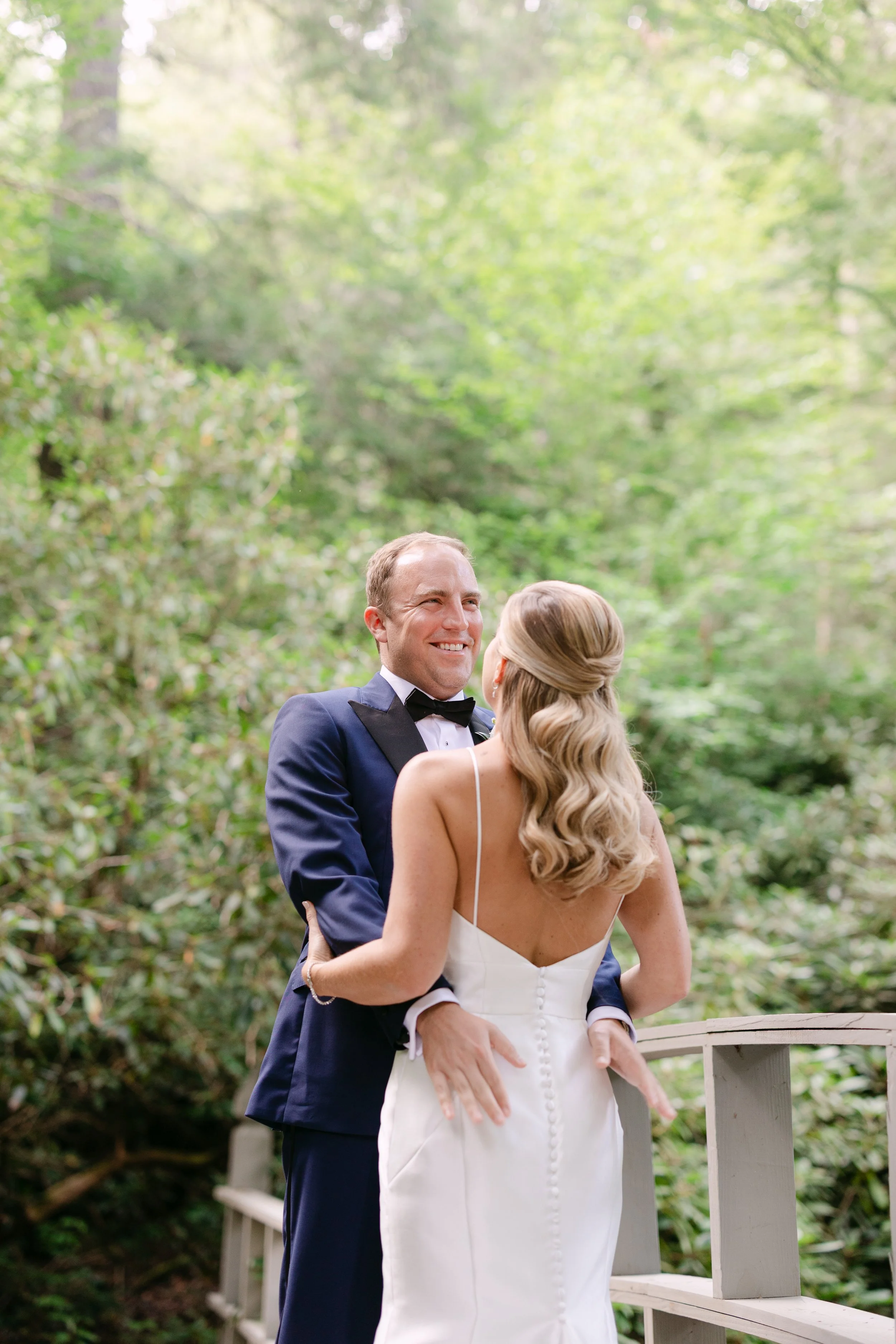 A couple in wedding attire sharing a joyful moment outdoors on a wooden bridge surrounded by lush green trees.
