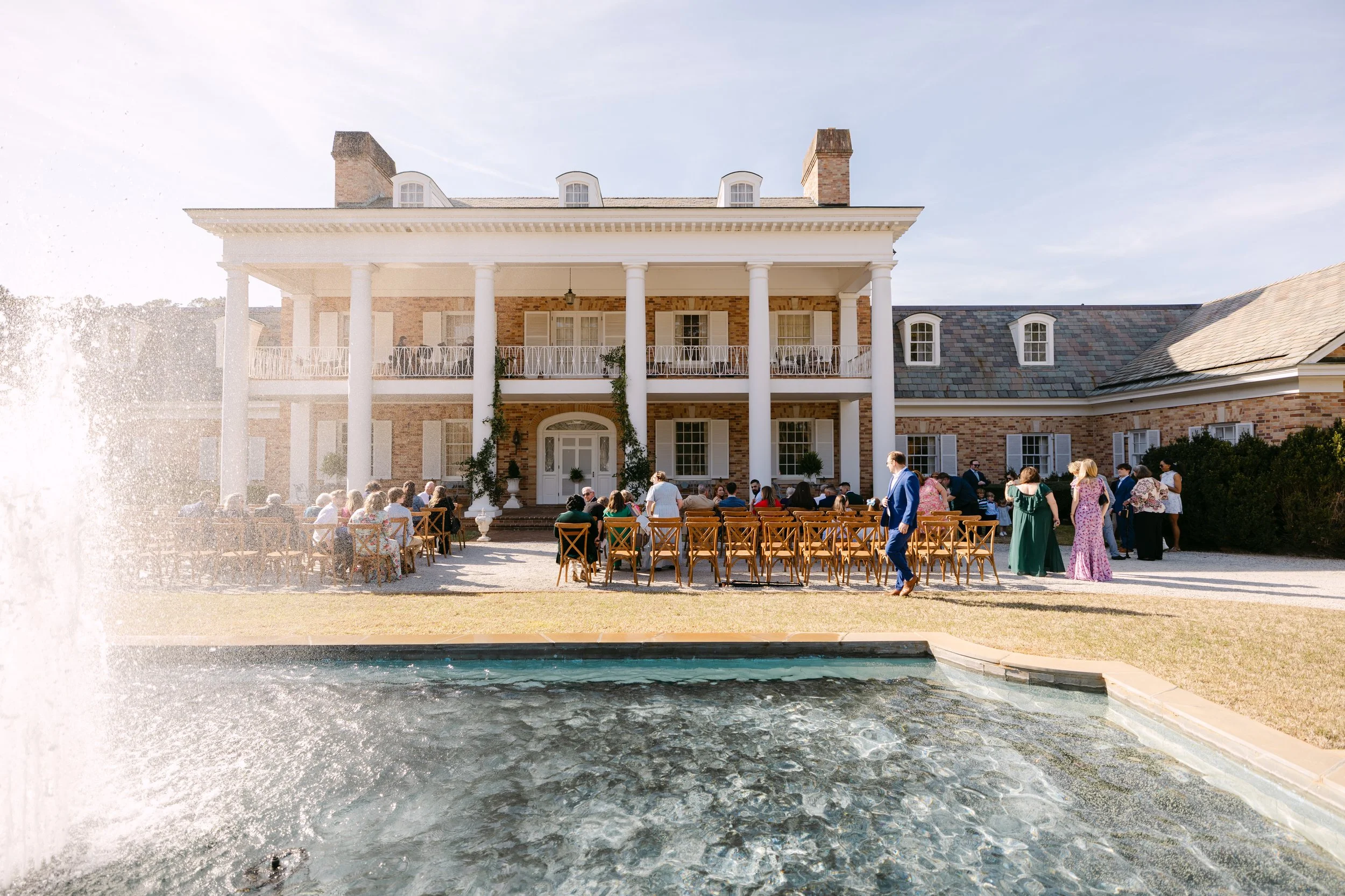 Guests gathering outside a large, brick mansion with a white-columned porch, attending a wedding ceremony on a sunny day.