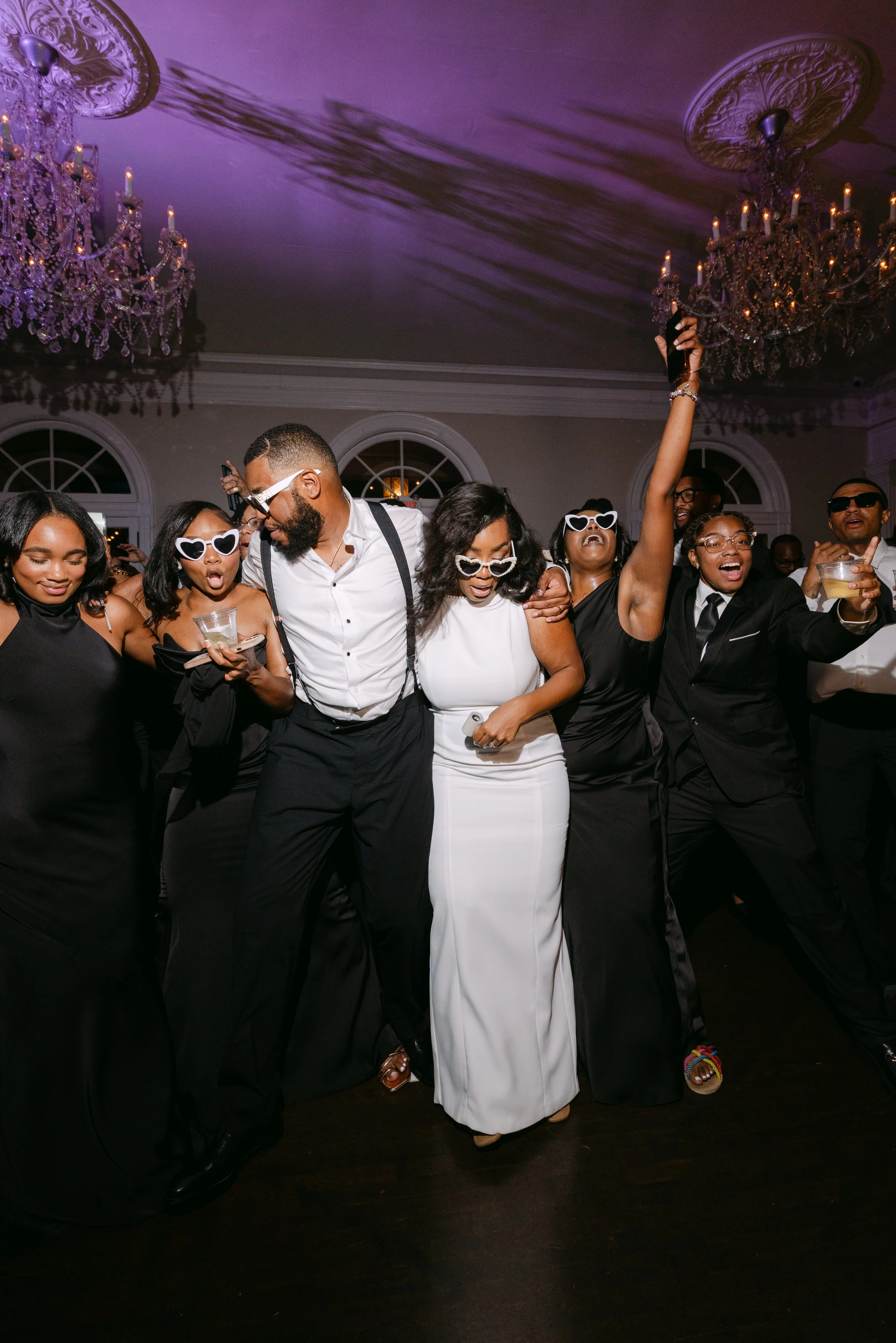 Group of people dancing and celebrating at a party, with some wearing heart-shaped sunglasses and holding drinks, in a room with chandeliers and elegant decor.
