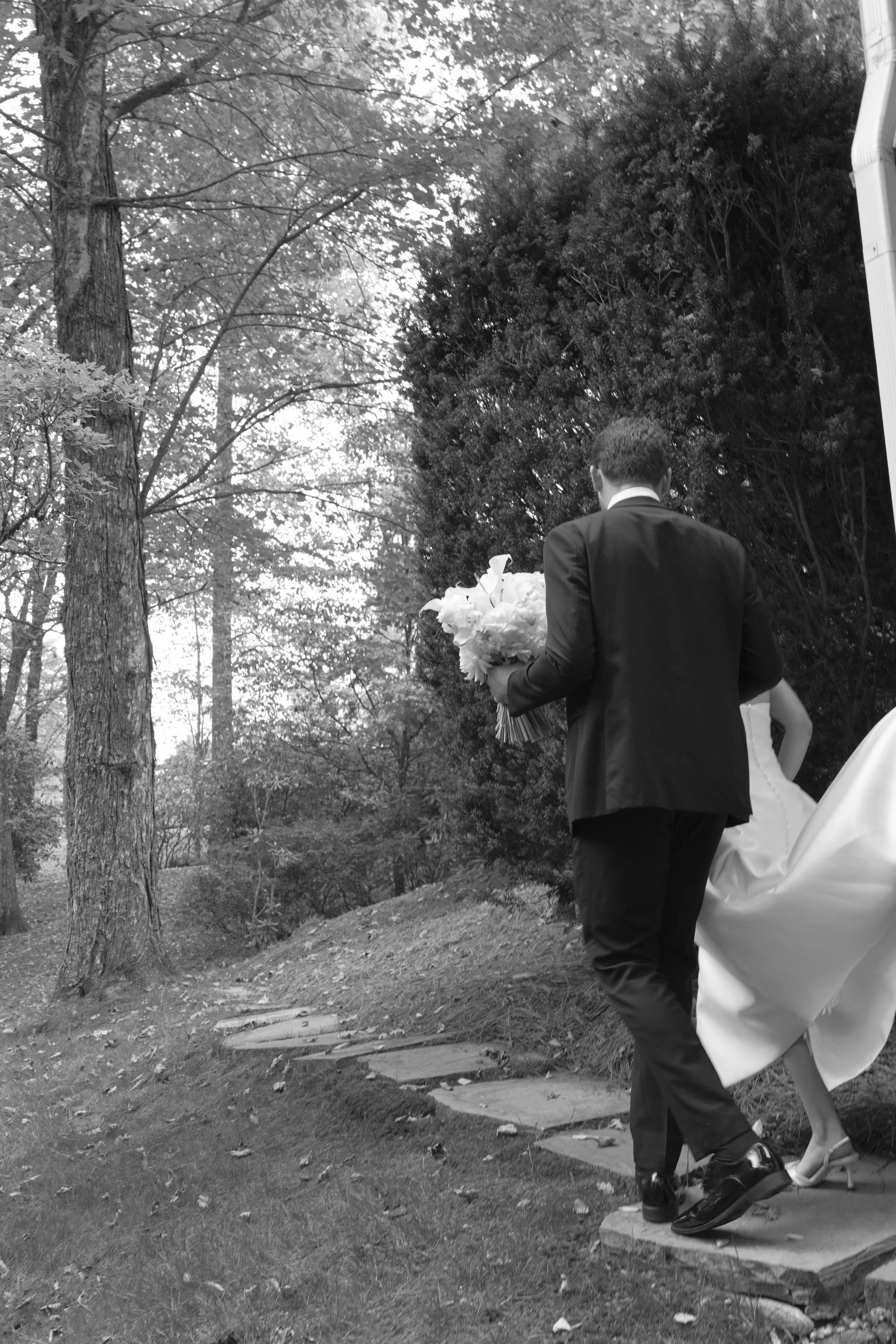 Bride and groom walking along a wooded garden path, with the groom carrying a bouquet.