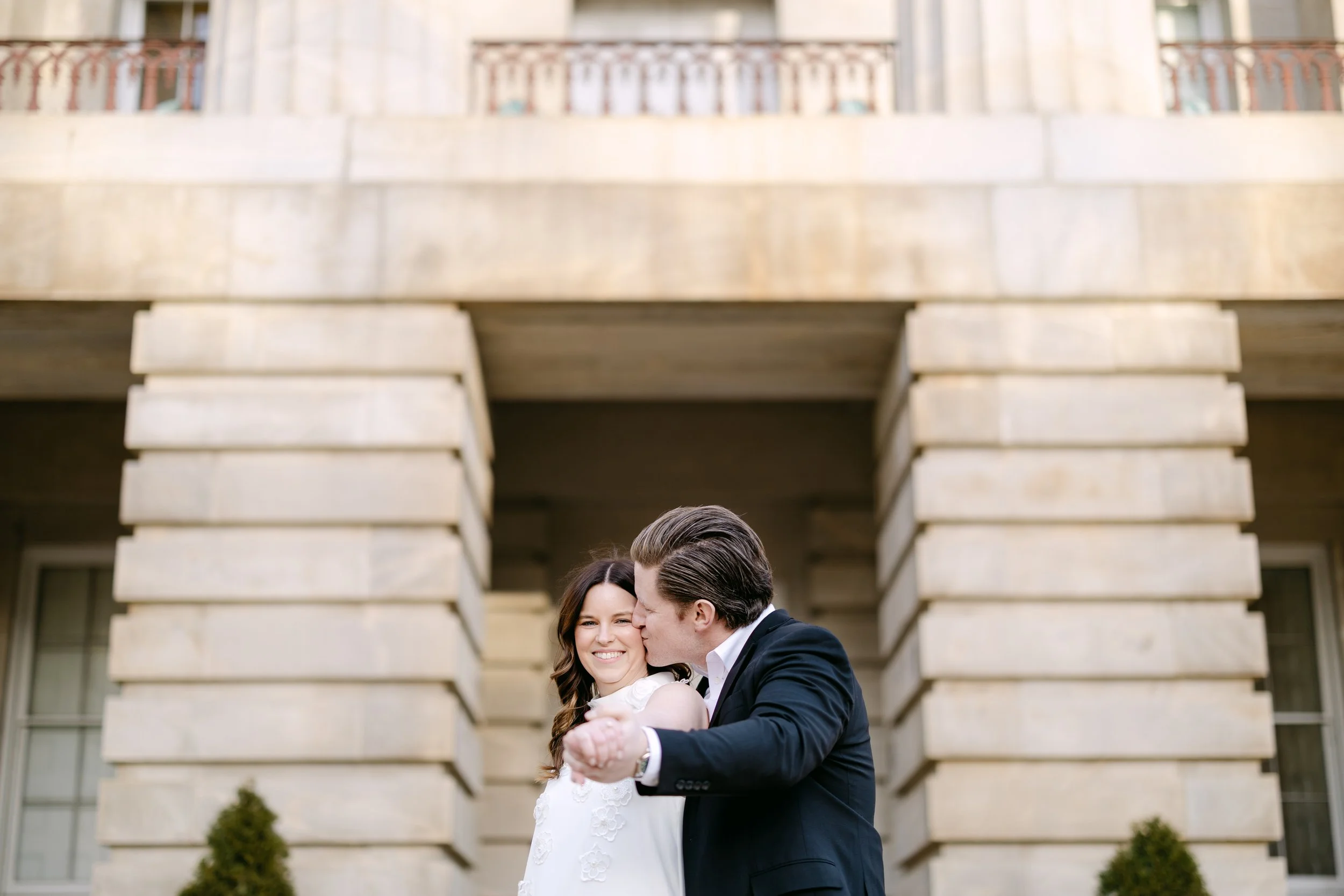 A couple dancing outdoors in front of a stone building, smiling and sharing a kiss.