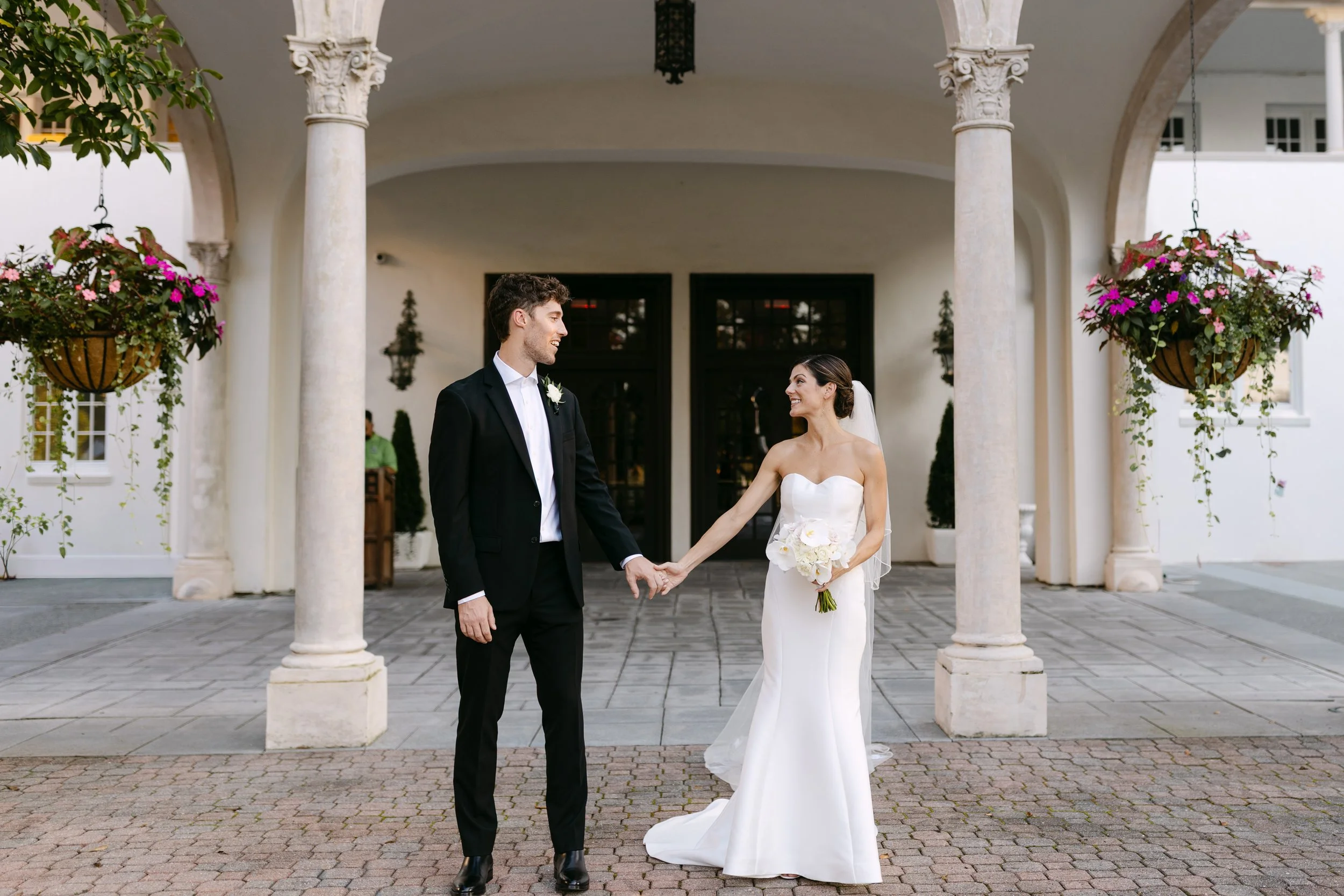 A bride and groom holding hands outside a wedding venue, smiling at each other.