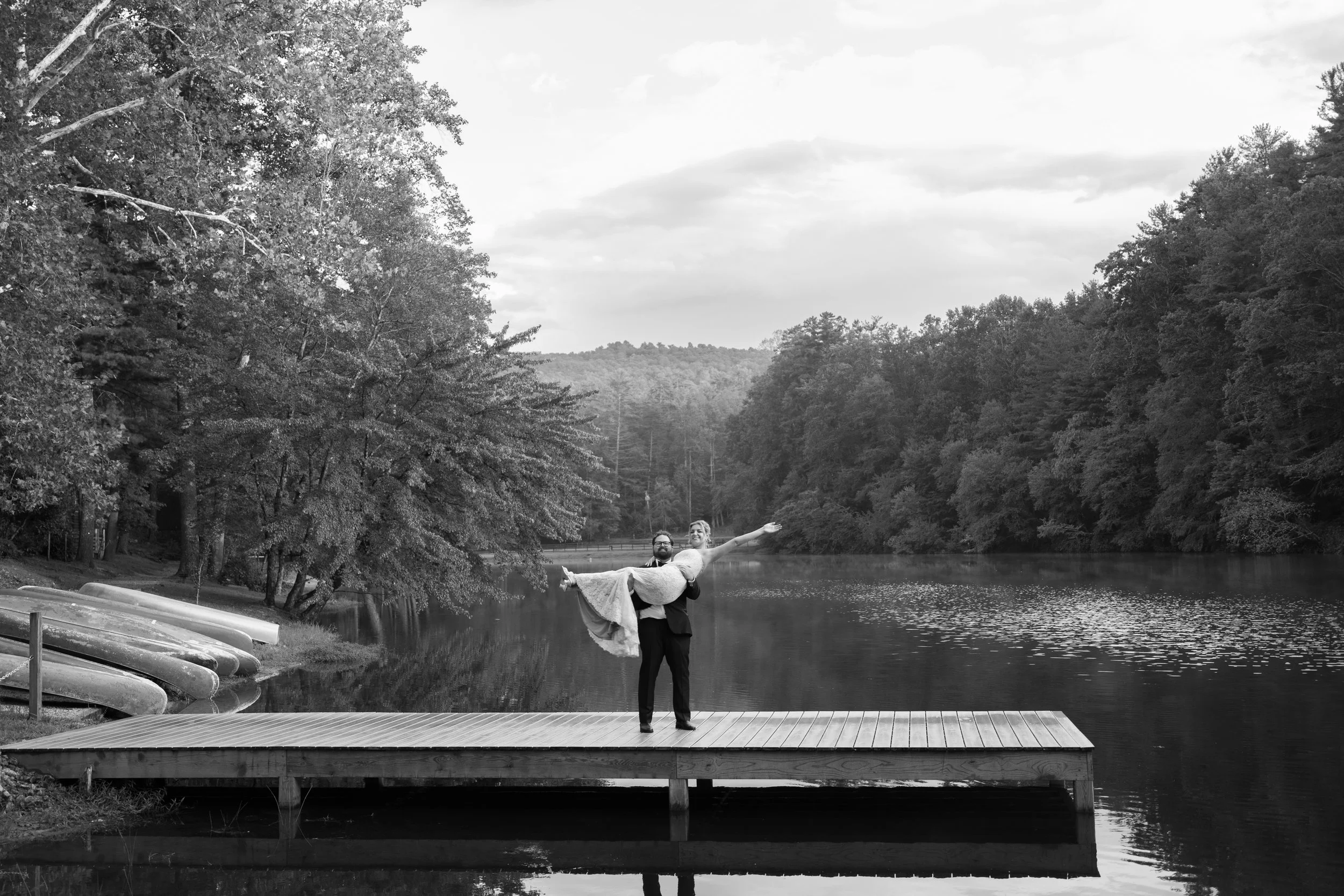 A couple stands on a dock by a lake, with the man holding the woman in his arms as she extends her arms and smiles, surrounded by trees and mountains in the background.