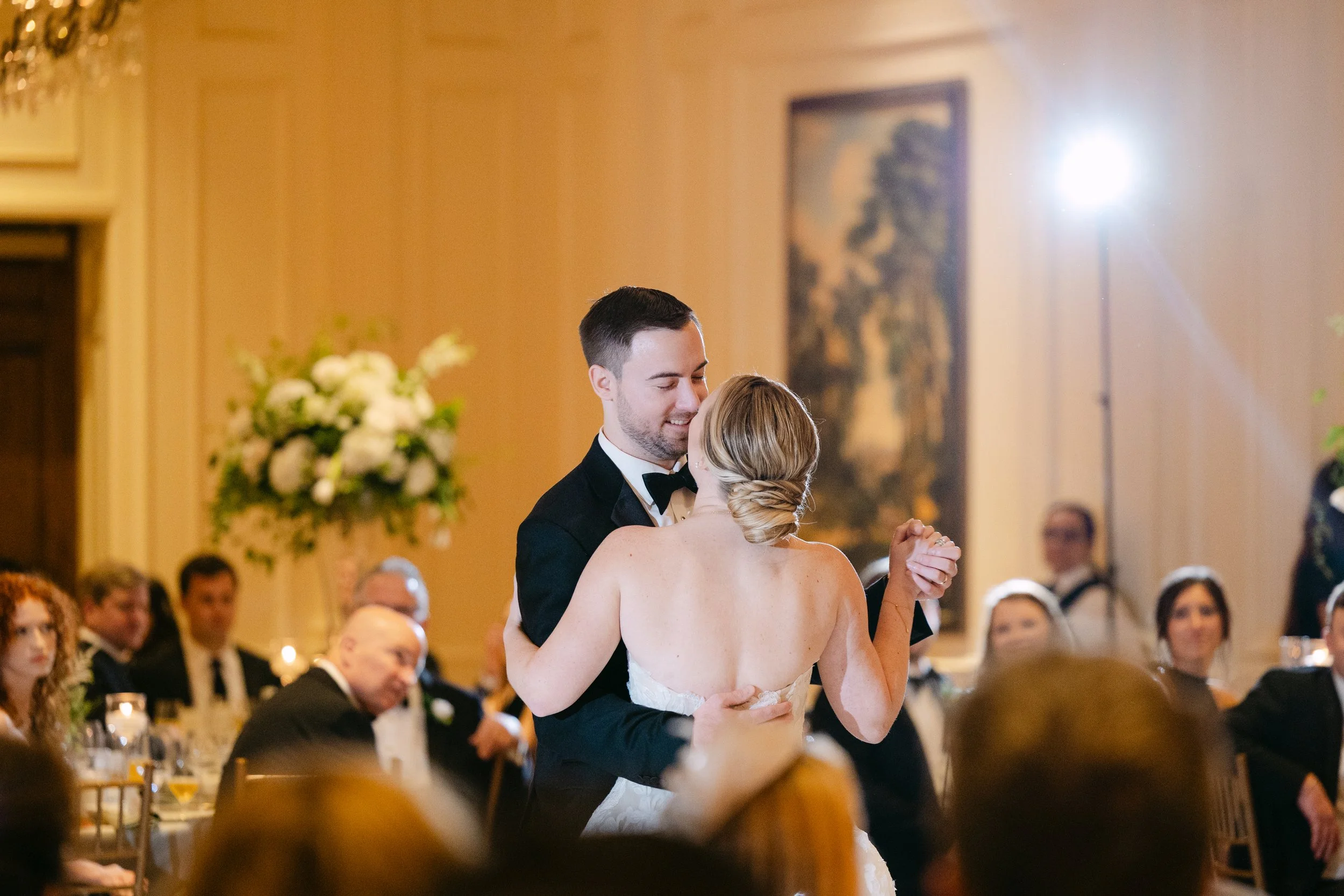 A bride and groom share a dance at their wedding reception, surrounded by seated guests in an elegant decorated ballroom.