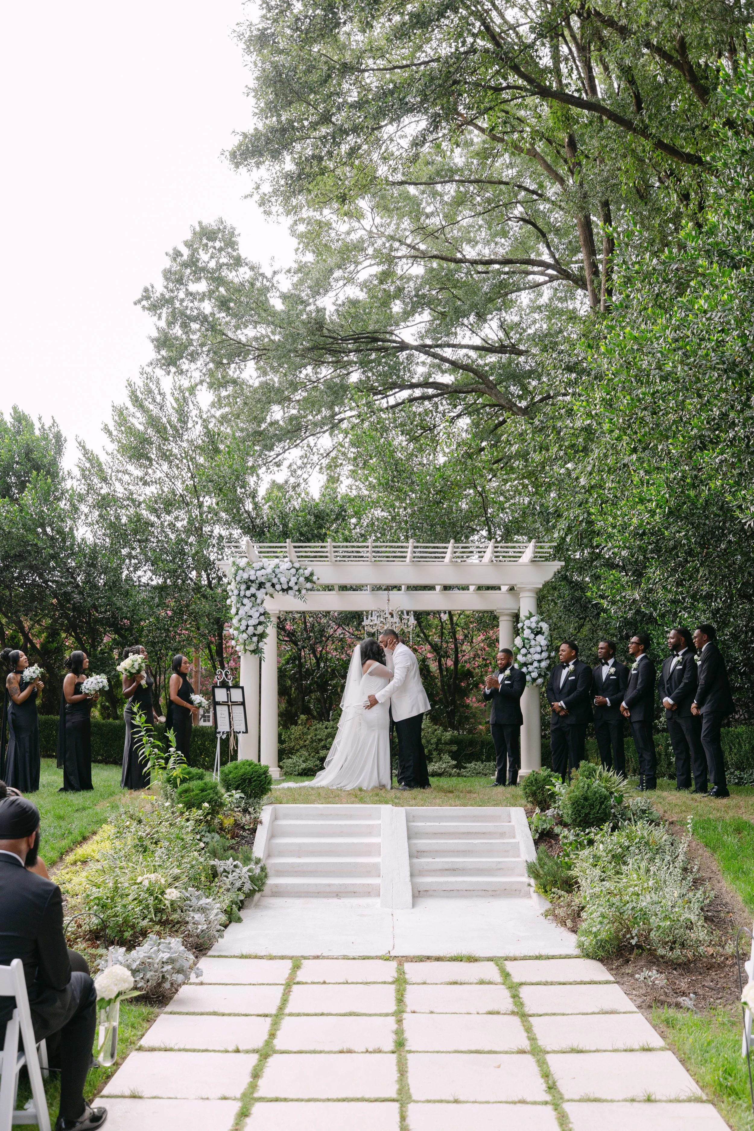 A wedding ceremony taking place outdoors under a white arbor decorated with white flowers, with a bride and groom kissing, surrounded by bridesmaids and groomsmen in formal attire, with guests seated nearby, in a lush garden setting.