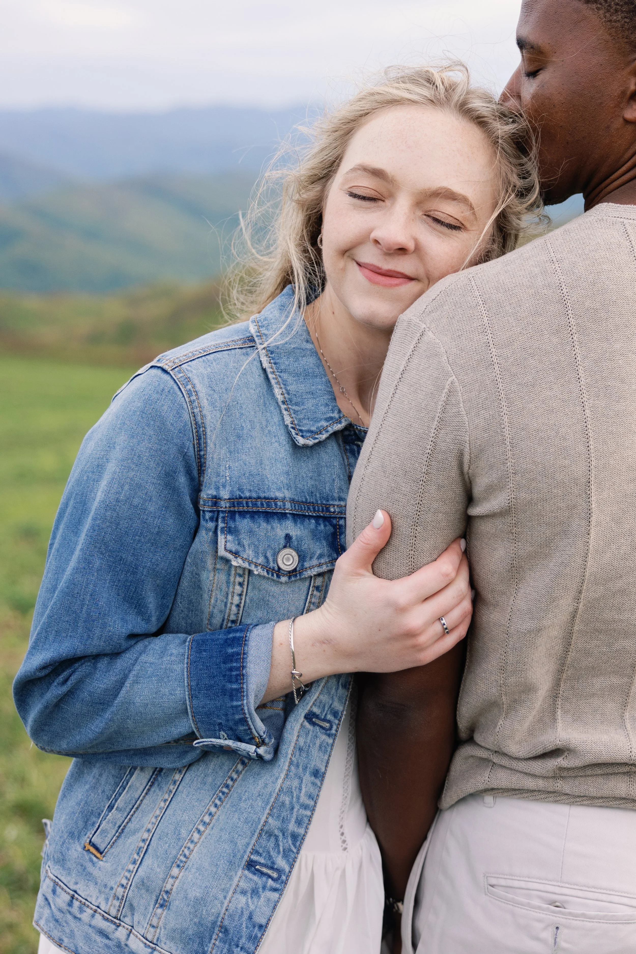 A woman with blonde hair and a denim jacket smiling with eyes closed, hugging a man with dark skin and a beige jacket, outdoors with a blurred green landscape and mountains in the background.