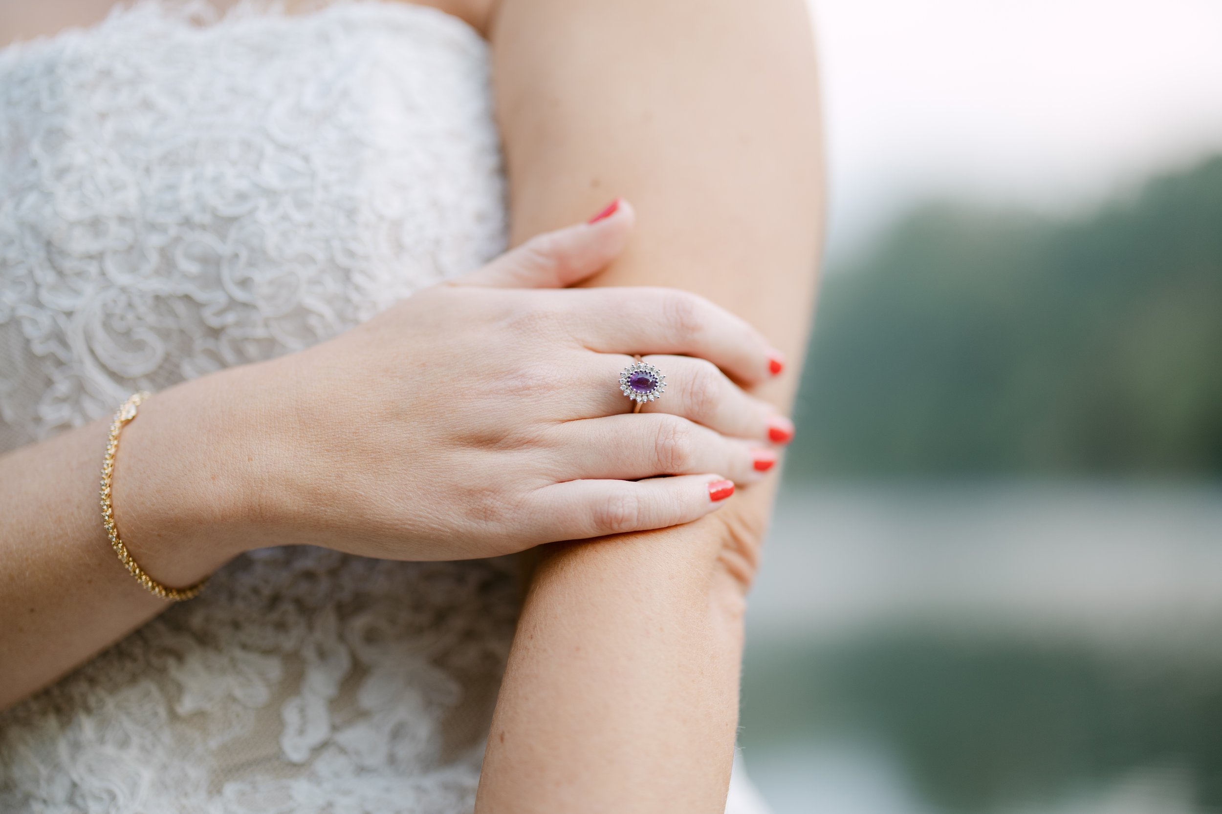 A woman's hand resting on her arm, showing a silver ring with a large purple gemstone surrounded by smaller diamonds, red nail polish, and a gold bracelet, with a lace dress in the background.