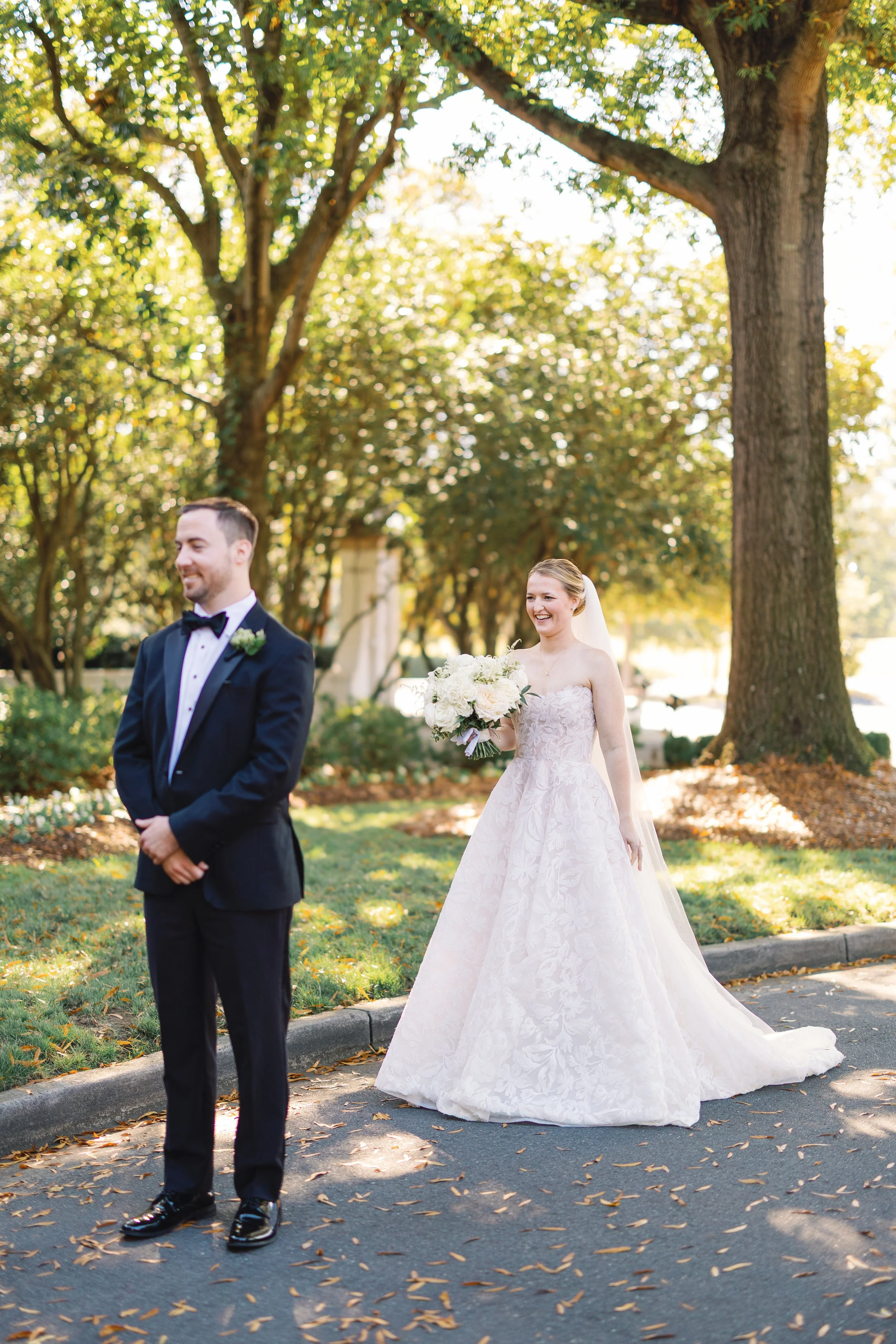 Bride in a white wedding gown holding a bouquet of white flowers stands behind a groom in a black tuxedo with a bow tie. They are outdoors on a sunny day with trees and fallen leaves on the ground.