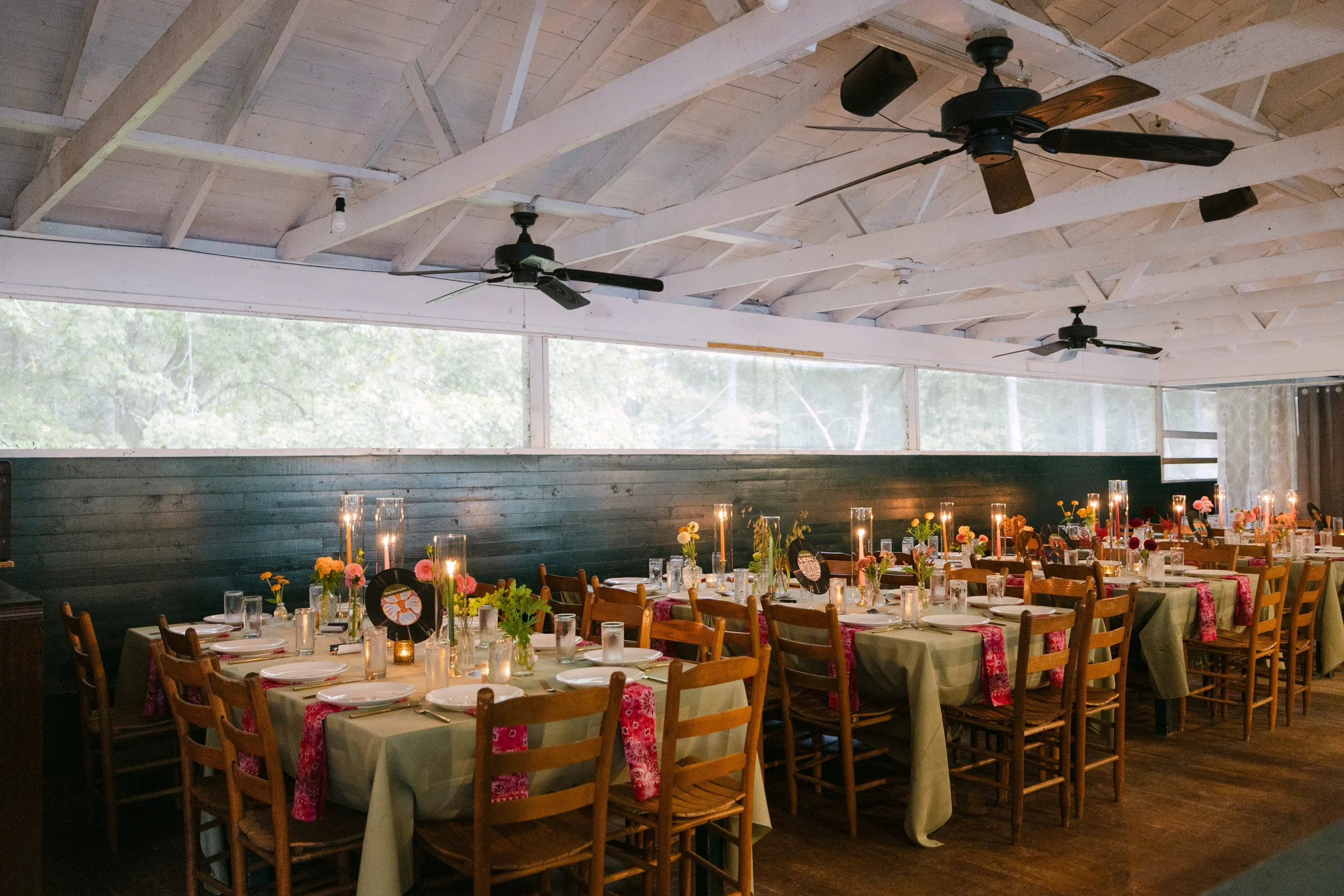 A decorated banquet hall with multiple tables set for a celebration, featuring floral centerpieces, candles, and place settings, under a wooden ceiling with ceiling fans.