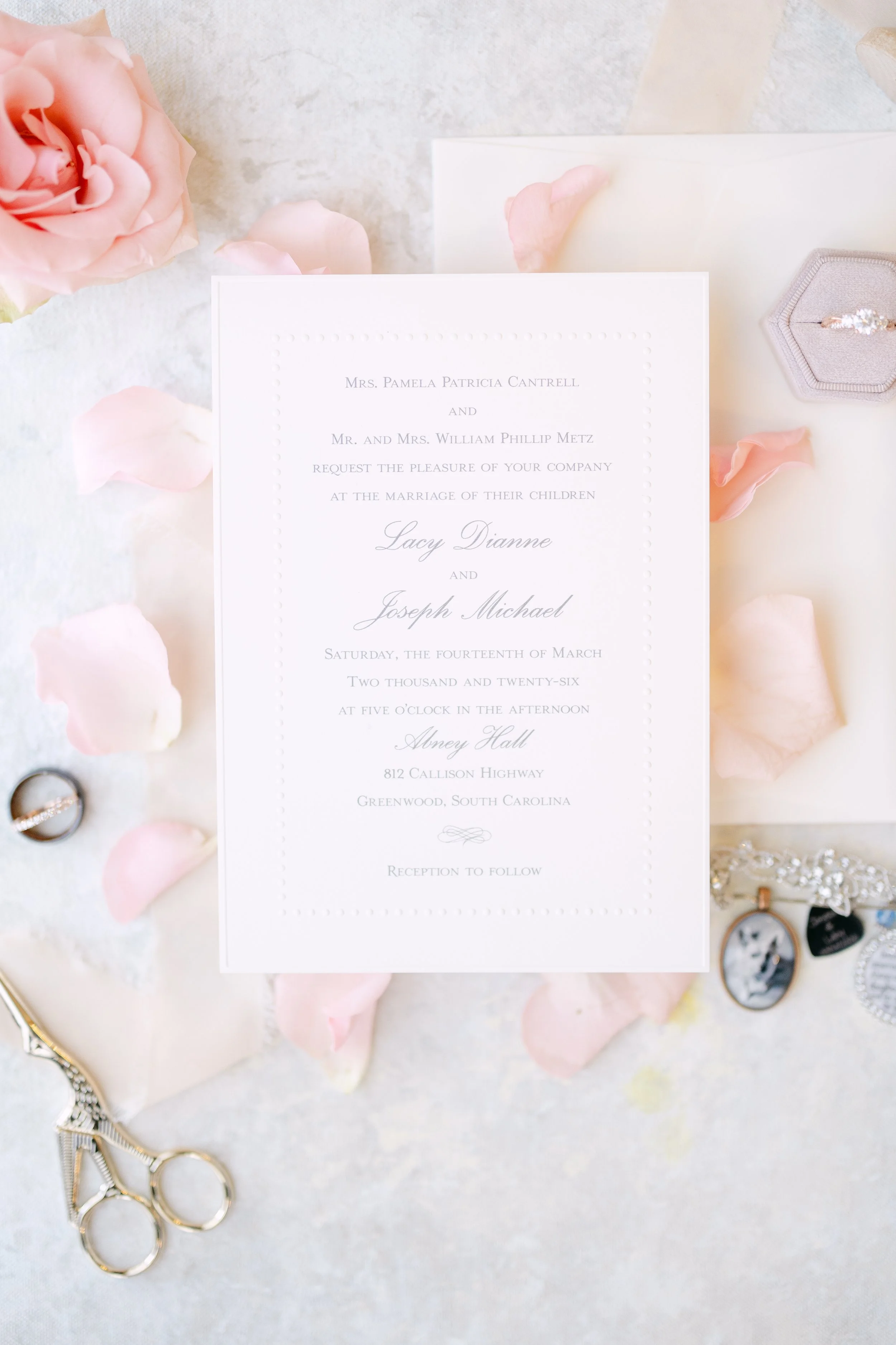 Wedding invitation on a light surface surrounded by pink flower petals, a wedding ring, a small pair of scissors, and jewelry including rings and a cameo photo pendant.