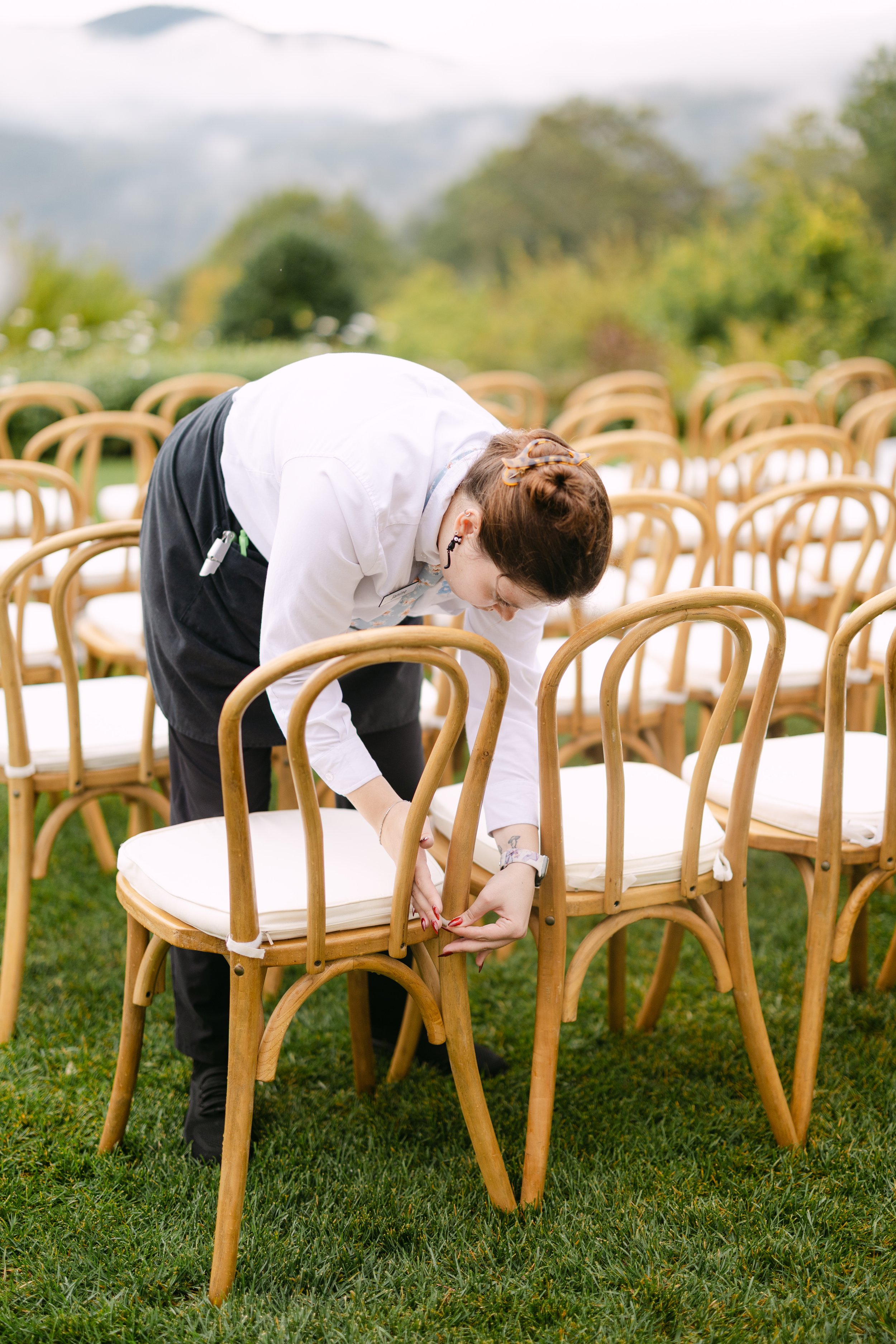 A person in formal attire adjusting a chair outdoors before an event setup with many other chairs on a grassy field, trees, and mountains in the background.