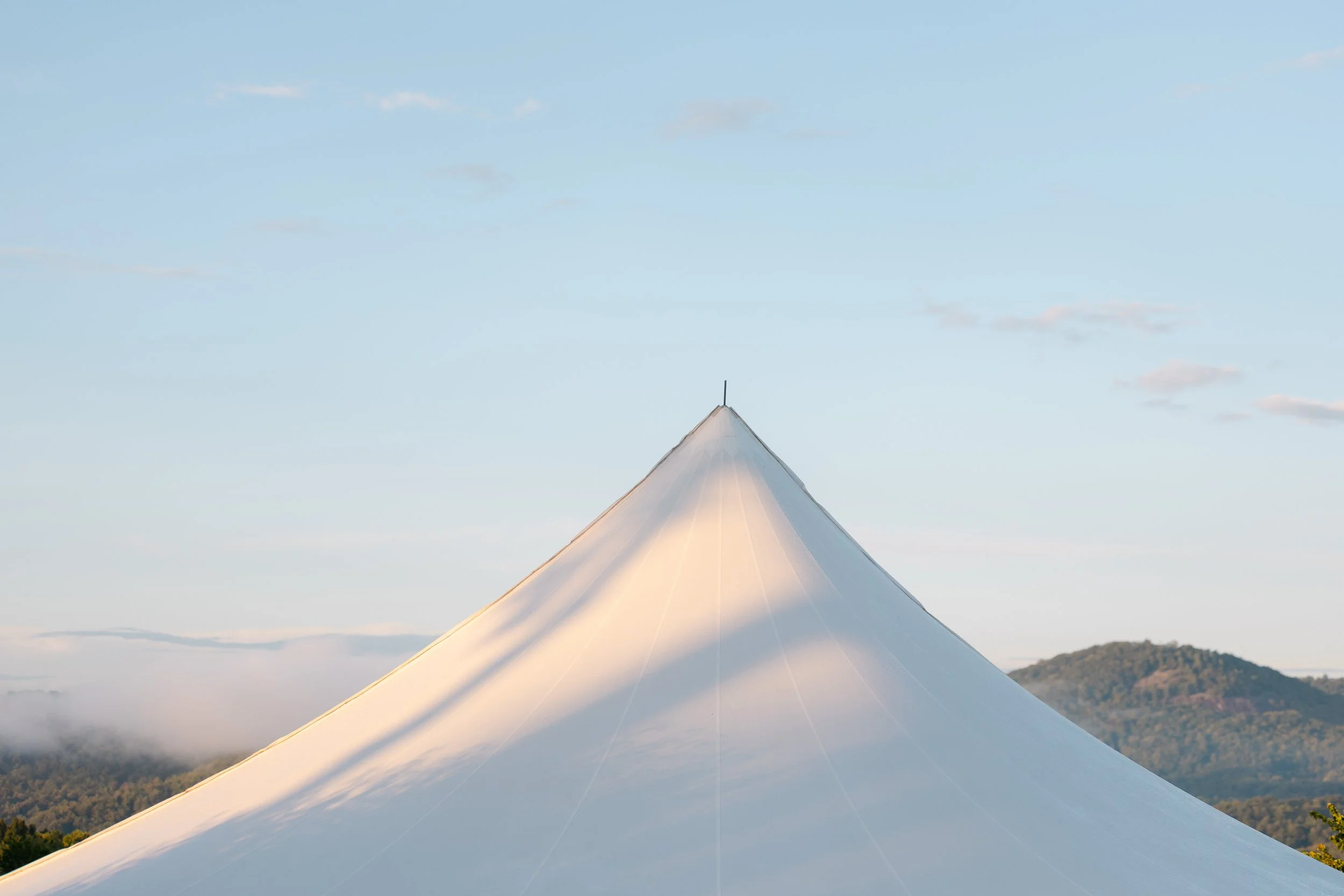 A large white tent with a pointed top, set against a backdrop of mountains and a partly cloudy sky.