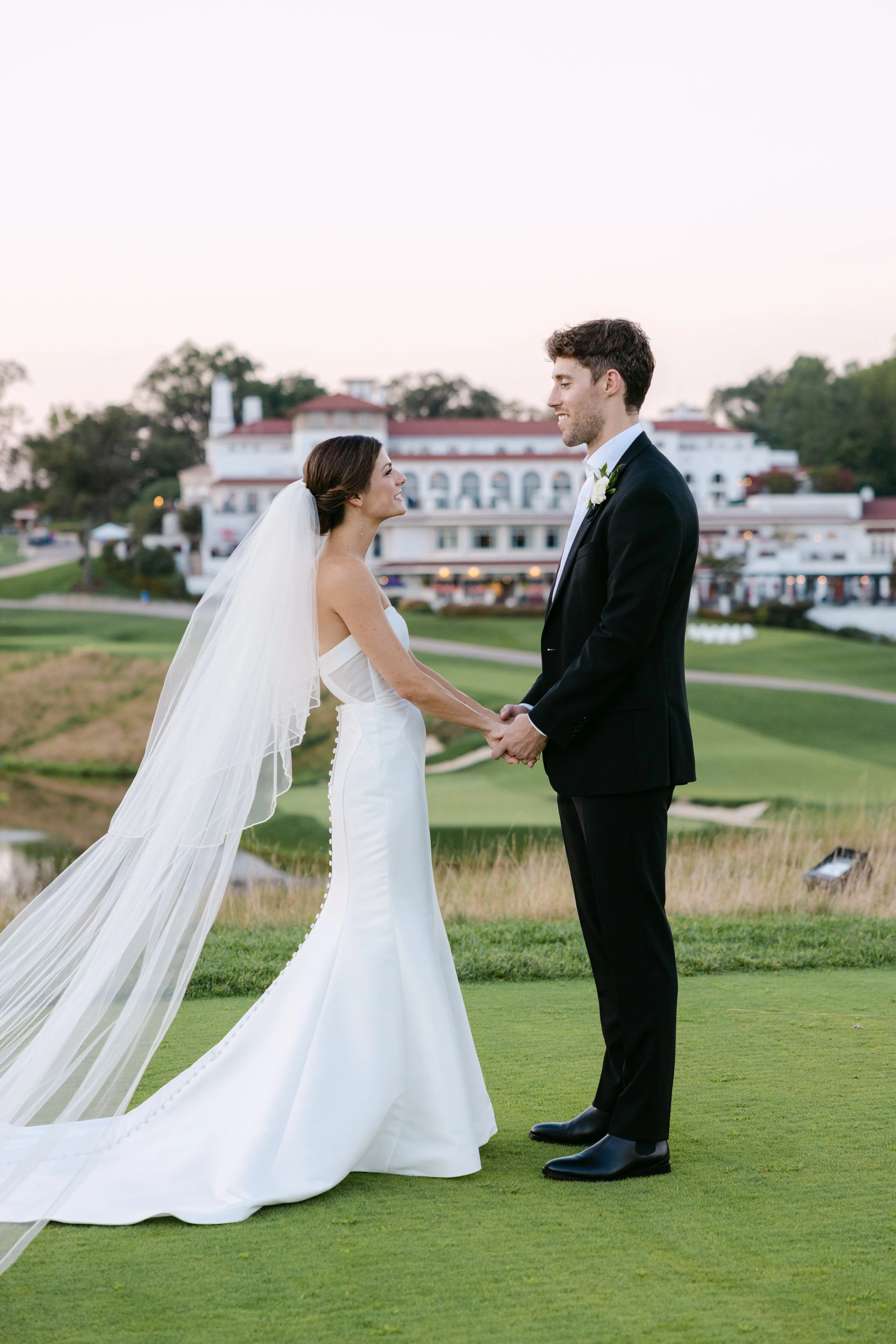 A bride and groom holding hands and smiling at each other on a golf course during their wedding, with a large white hotel building in the background at sunset.