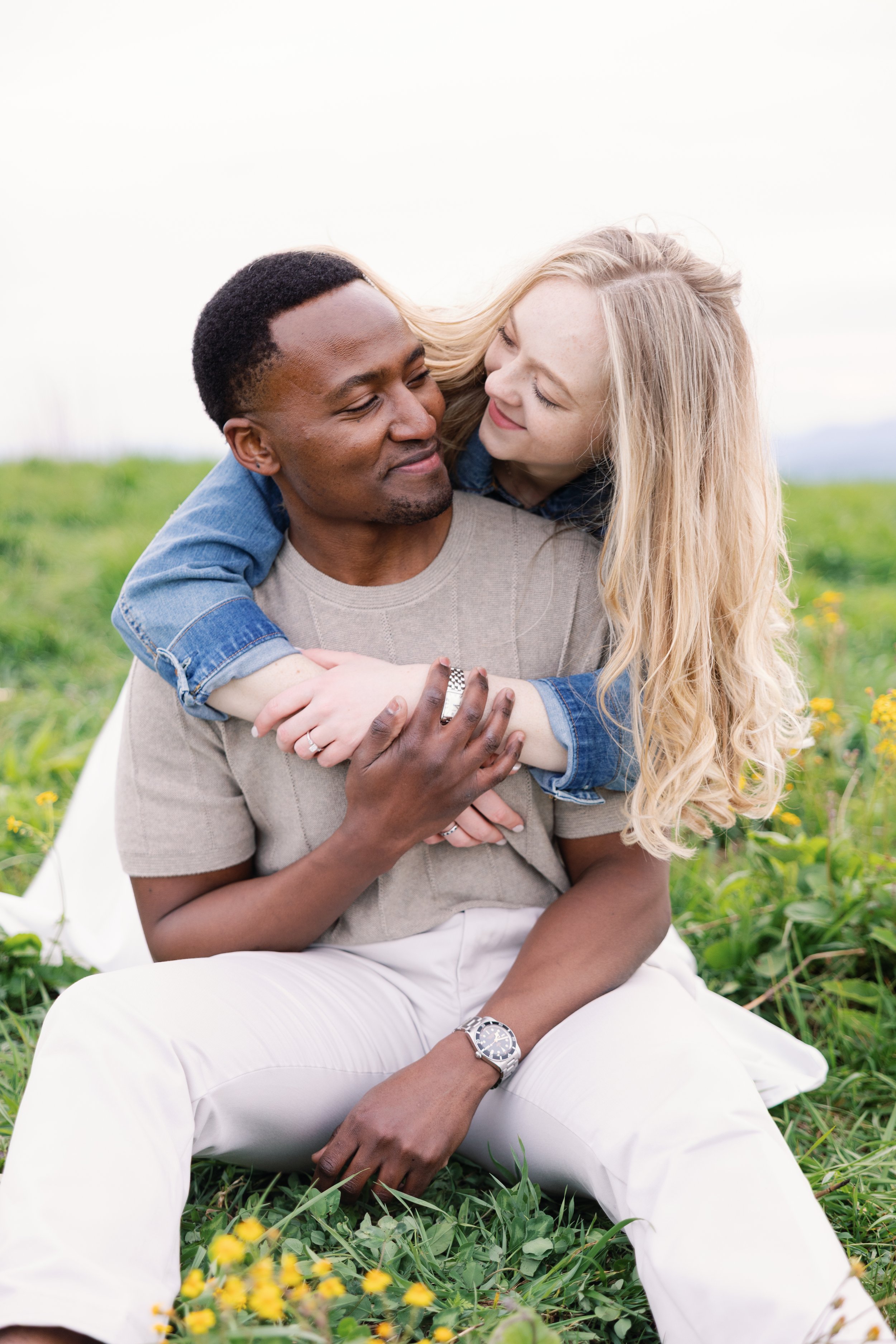 A black man and a white woman sharing a joyful moment outdoors, with the woman embracing the man from behind on a sunny day in a grassy field.