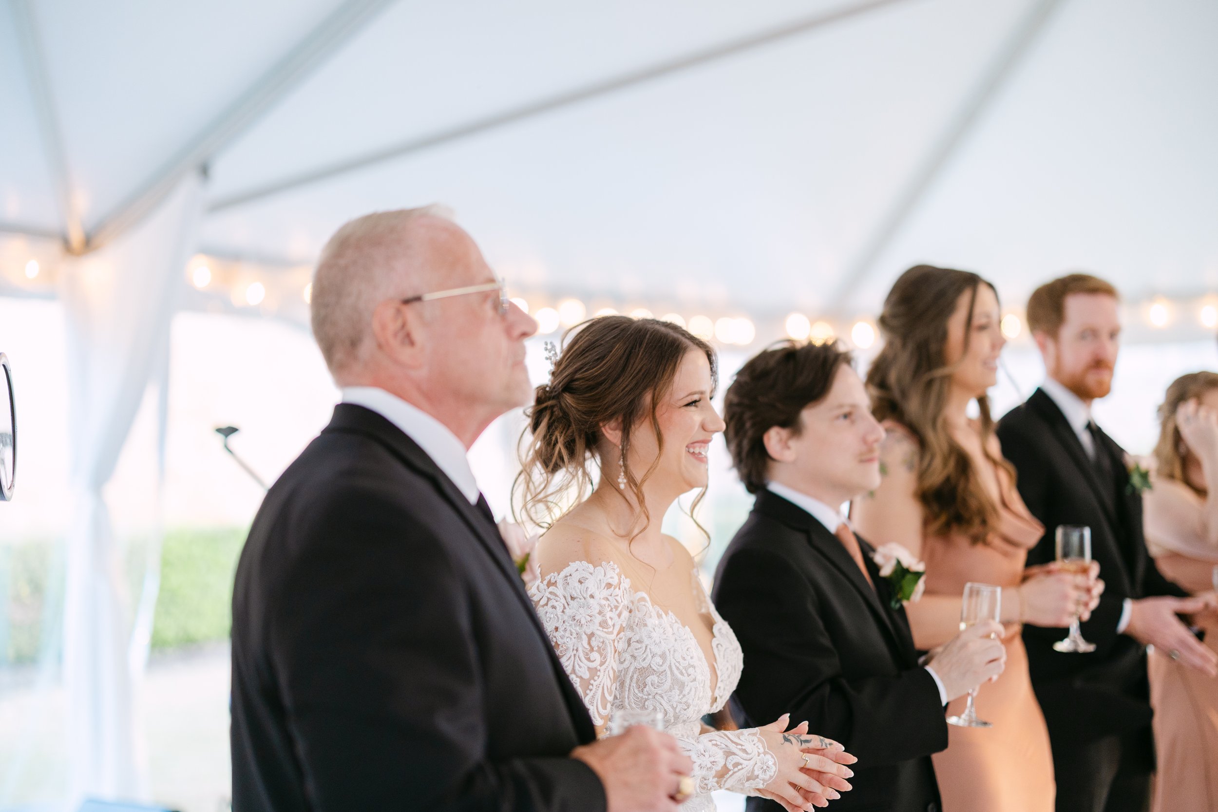Group of people at a wedding reception, standing under a white tent, dressed in formal attire, holding champagne glasses, smiling and celebrating.