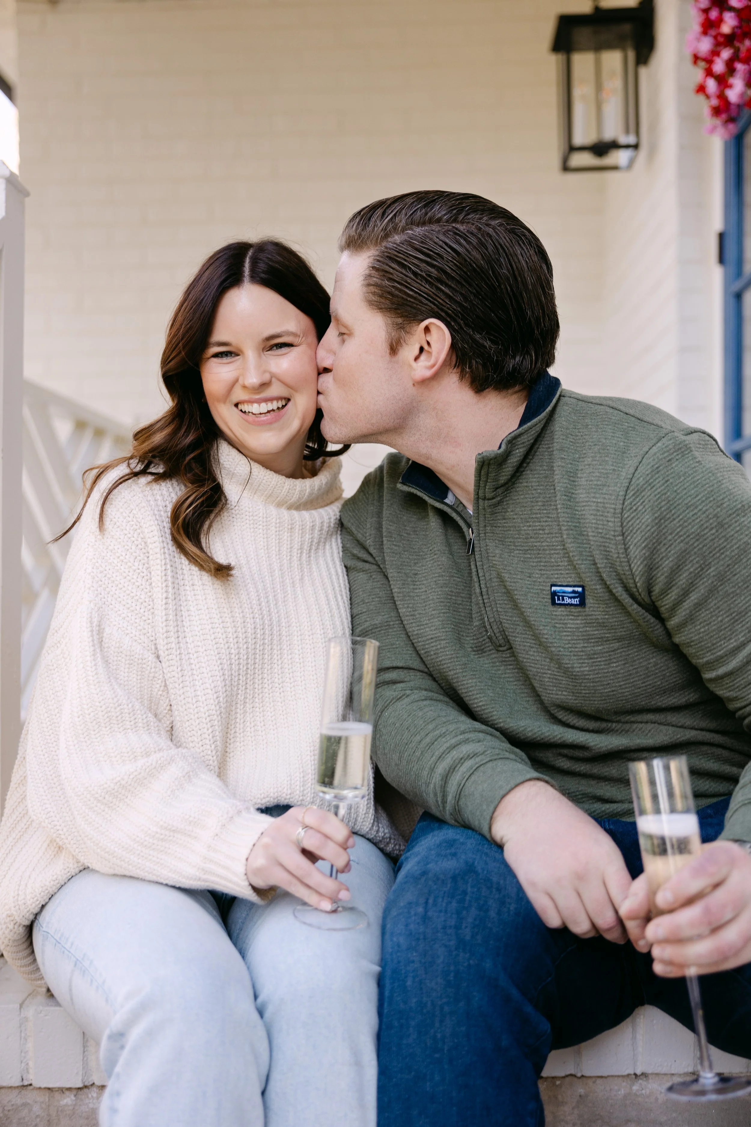 A man kisses a woman on the cheek while sitting outside with champagne glasses, both smiling.