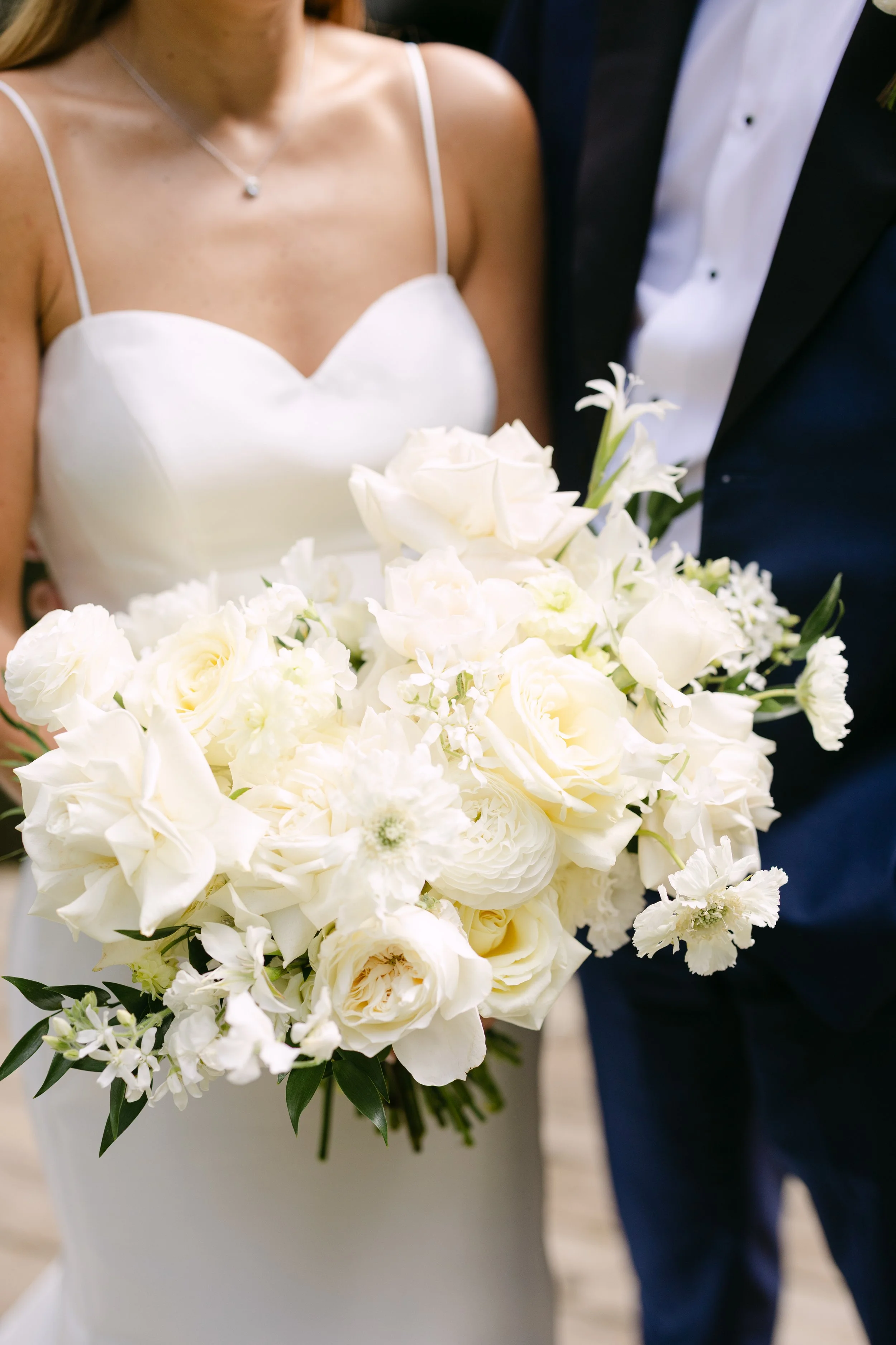 A bride holding a large bouquet of white flowers at her wedding, next to a groom in a tuxedo.