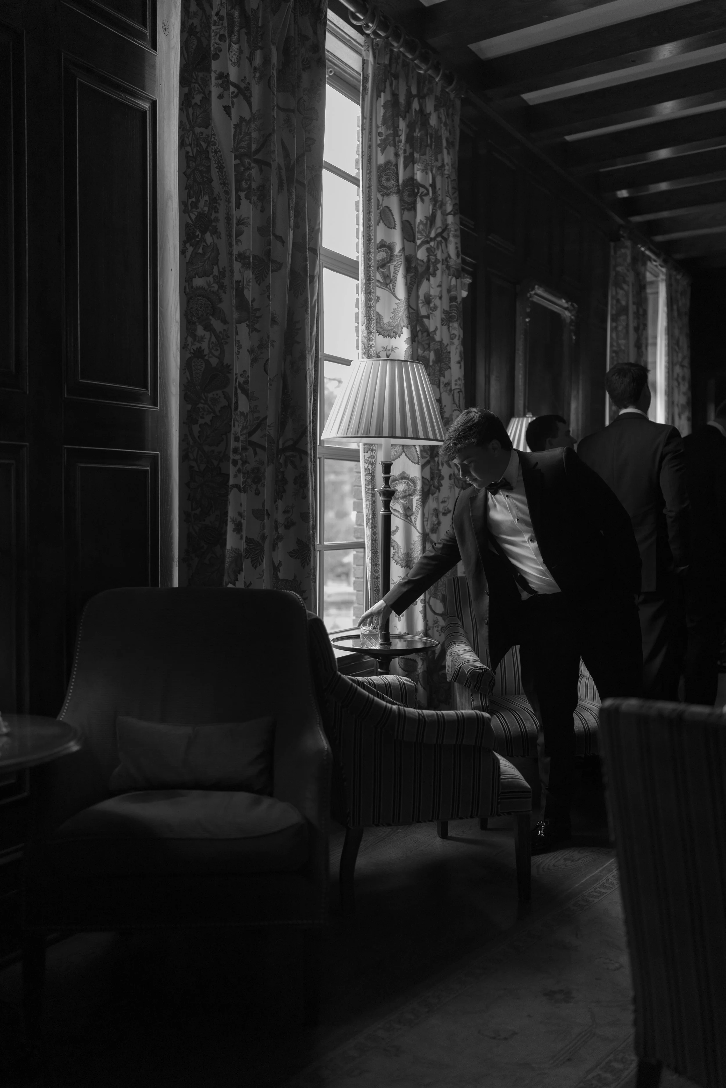 Black-and-white scene of a tuxedoed man reaching for a drink in an elegant room with floral curtains, upholstered chairs, and other formally dressed guests.