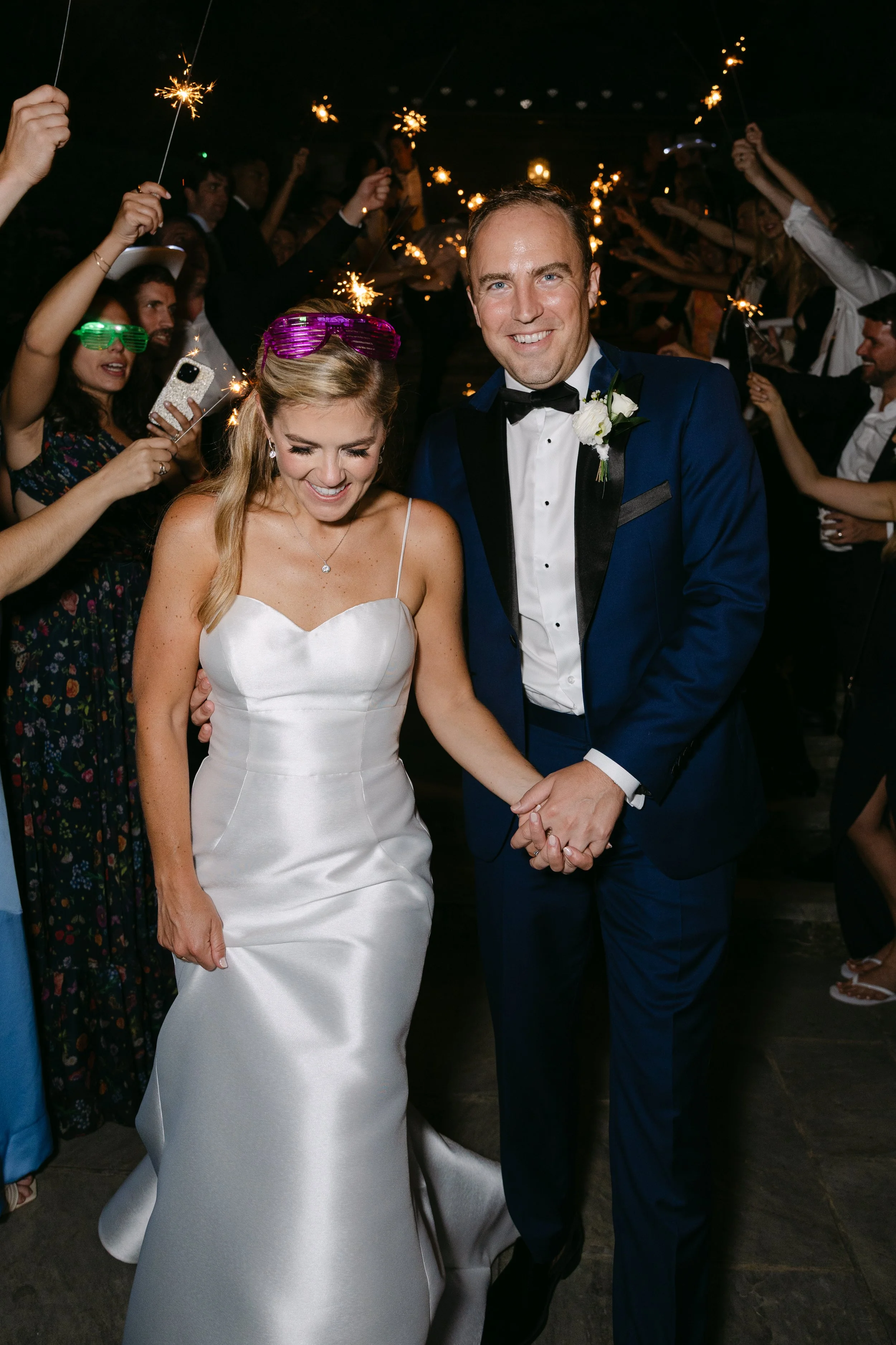 A newlywed couple dressed in wedding attire holding hands, surrounded by guests holding sparklers at night, celebrating their wedding.