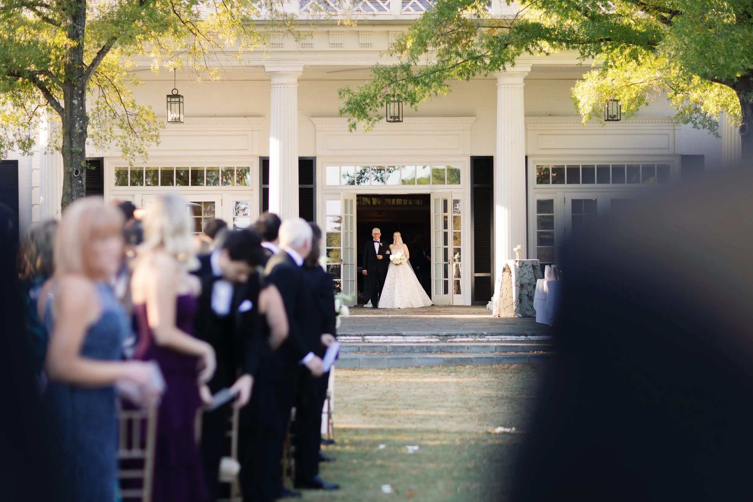 A bride walking down the aisle with her father at an outdoor wedding ceremony, with guests seated on either side and a white building with columns in the background.