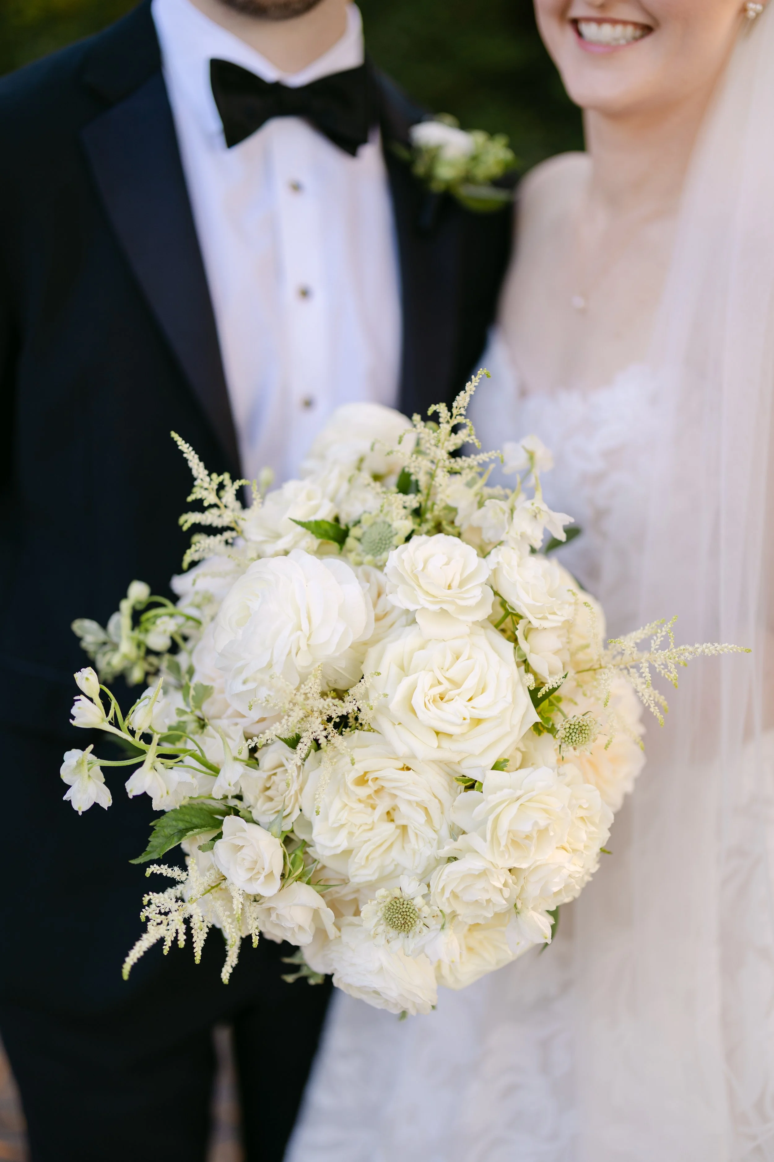 A bride and groom holding a large white floral bouquet outdoors, smiling and dressed in wedding attire, with the groom in a tuxedo and the bride in a wedding dress.