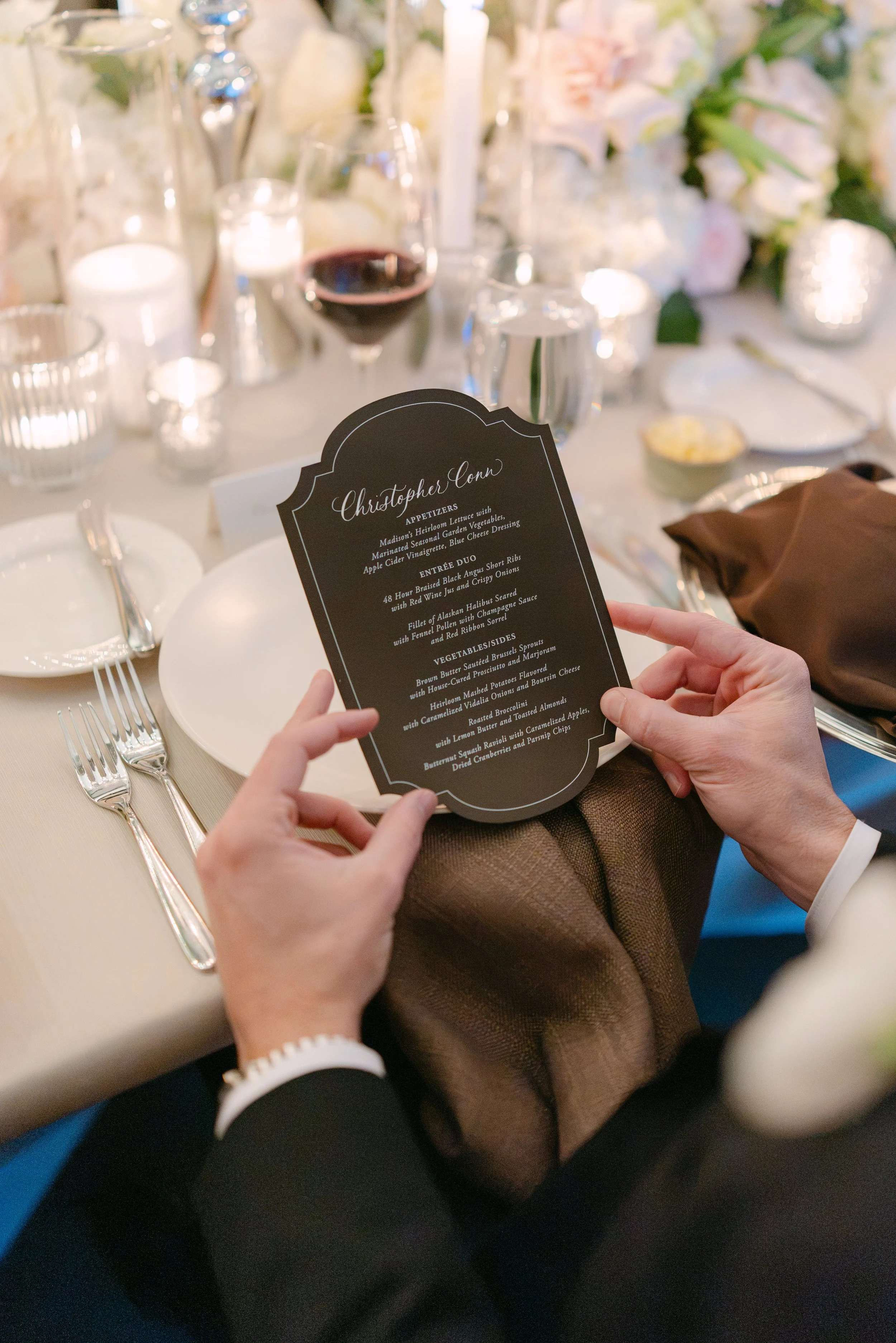 Elegant wedding reception table with a printed dinner menu, wine glasses, candles, place settings, and guests’ hands